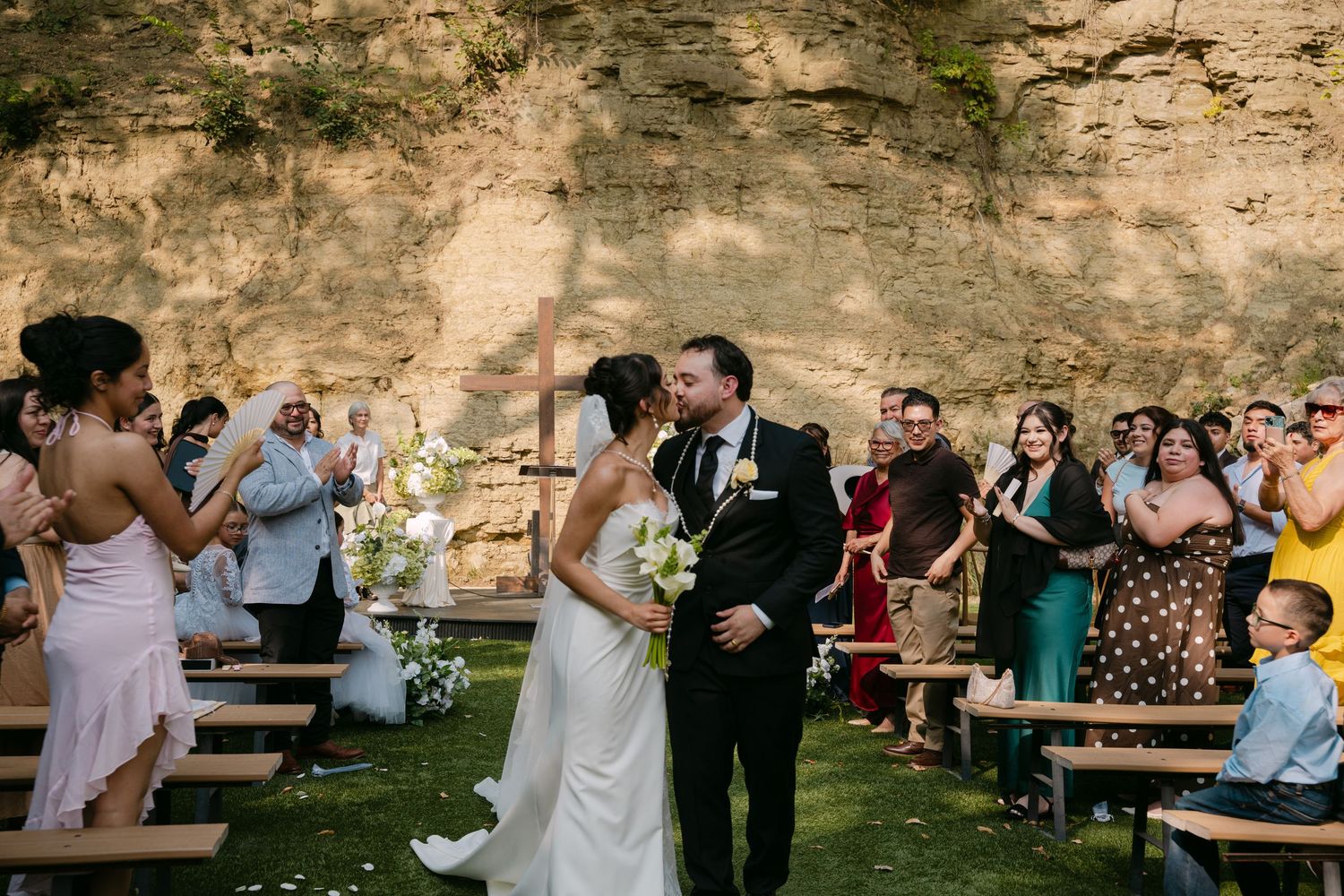 Newlyweds walk down the aisle as guests cheer at outdoor wedding ceremony near stone wall.