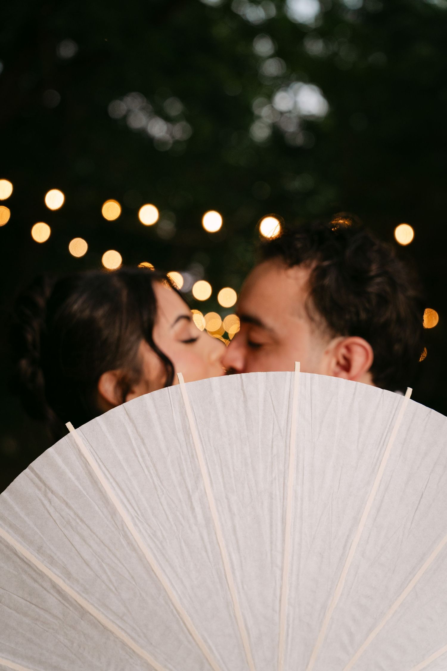 Bride and groom share a kiss hidden behind a white paper parasol with bokeh string lights above.