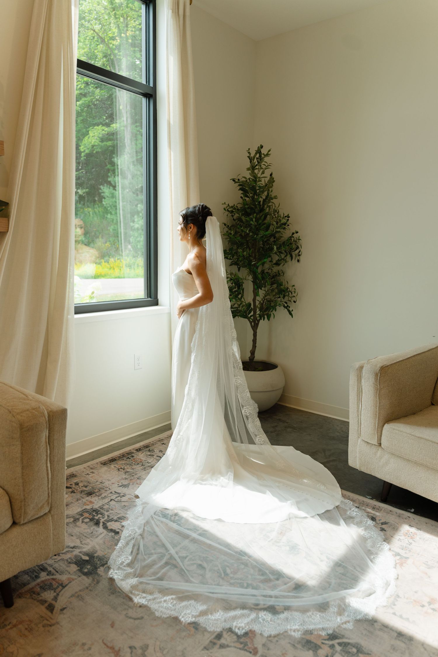 Bride in a white gown with long lace veil standing by a tall window in a bright room.