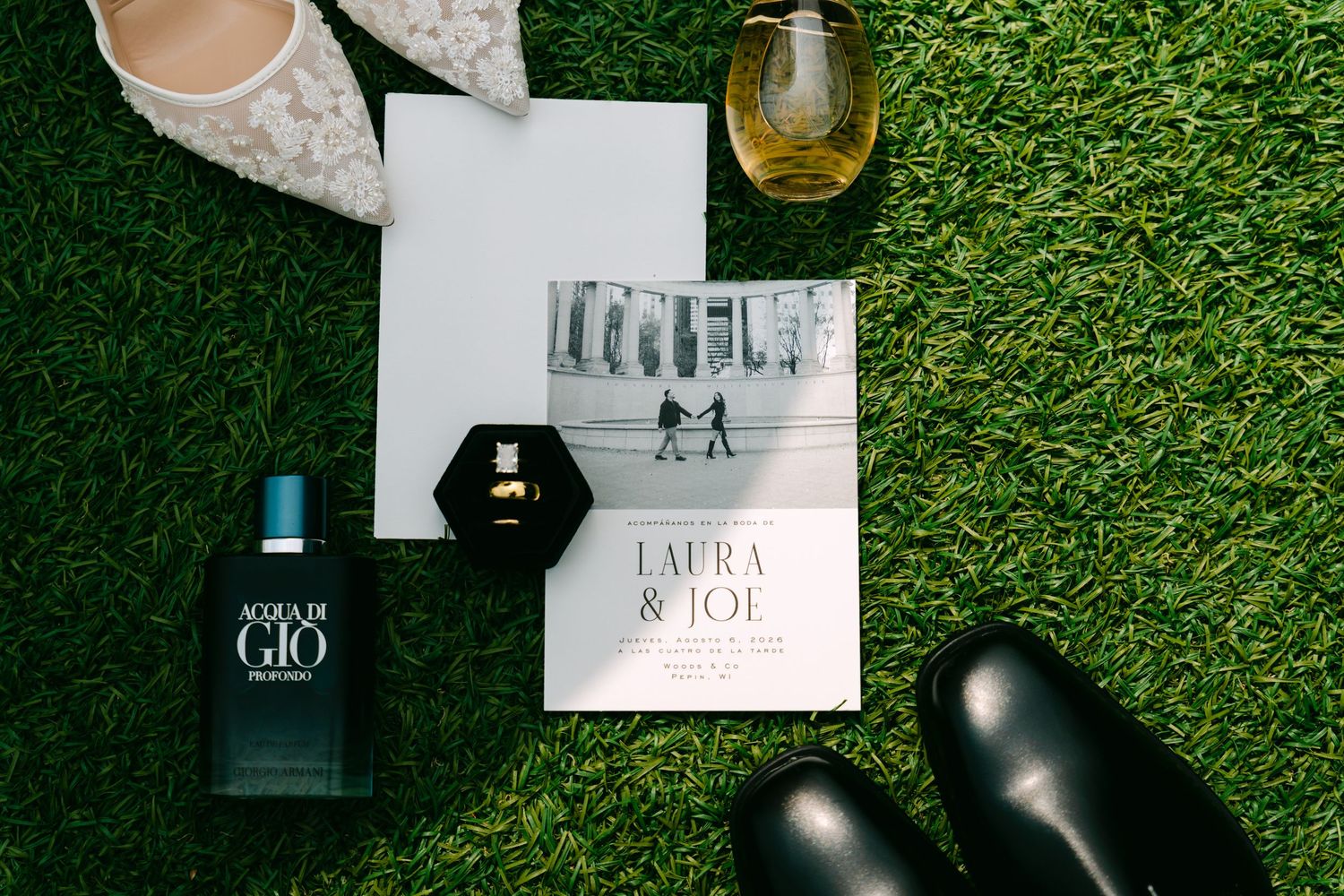 Wedding flat lay with Laura and Joe invitation, Acqua di Gio cologne, rings, heels, and boots on grass.