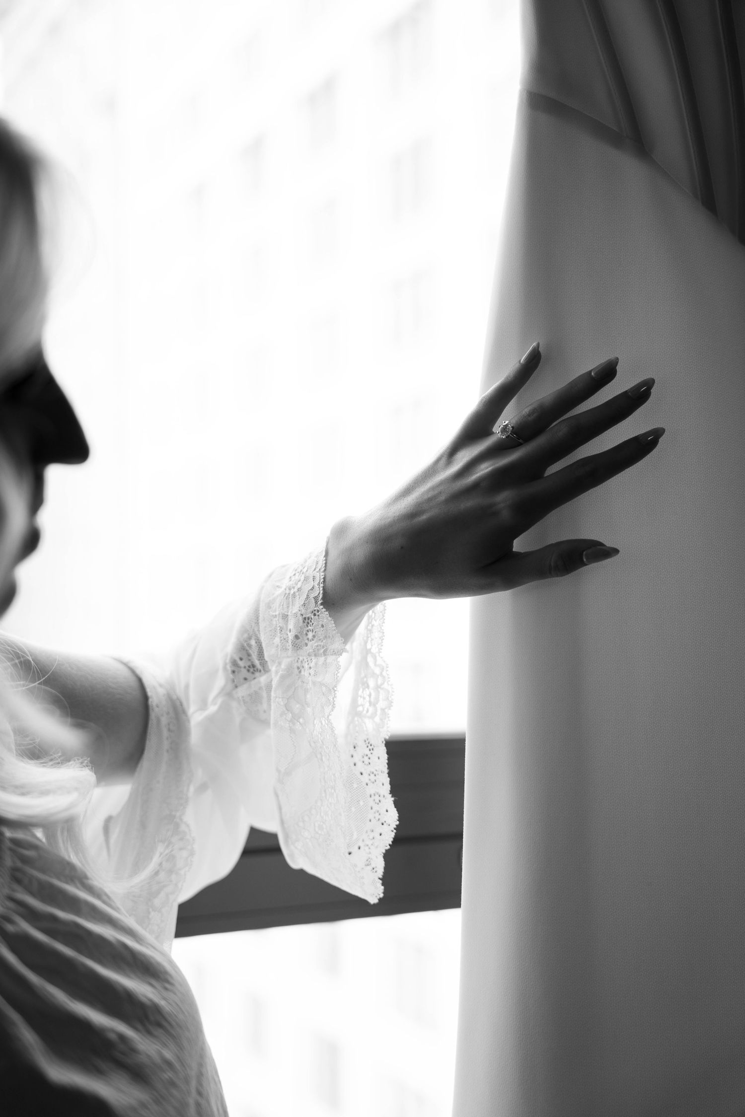Bride in white lace robe showing engagement ring while standing by bright window on wedding day.