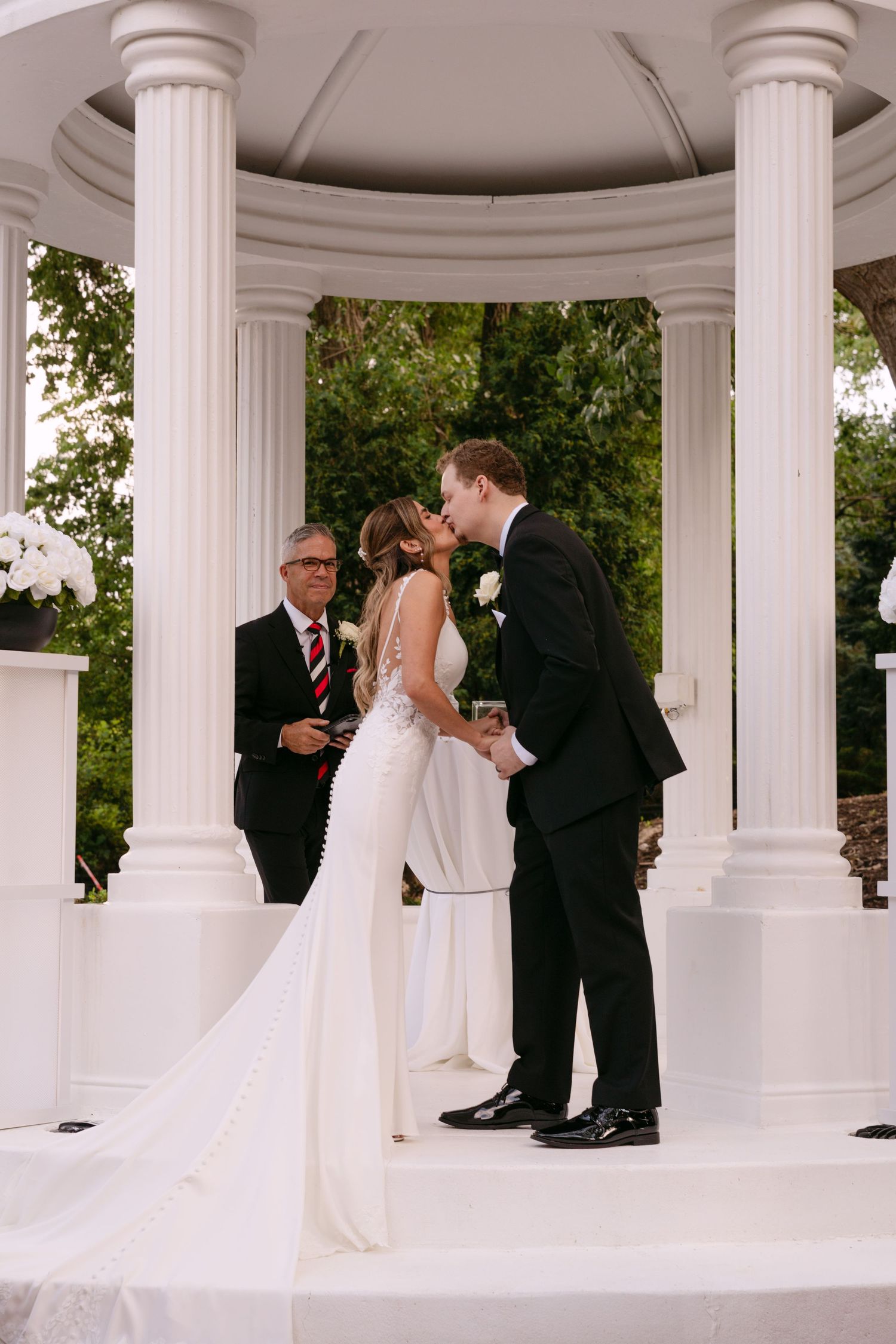 Bride and groom share first kiss at elegant white gazebo wedding ceremony with officiant and floral arrangements.