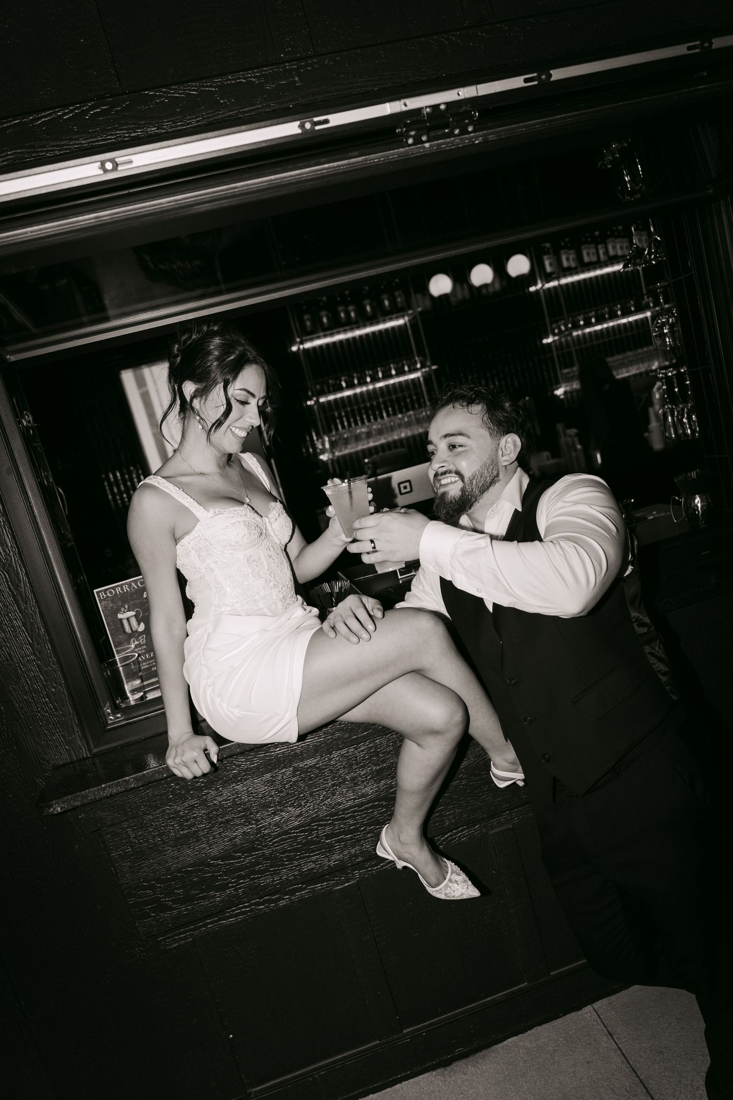 Newlyweds clink glasses at a bar counter in a classic black and white wedding portrait.