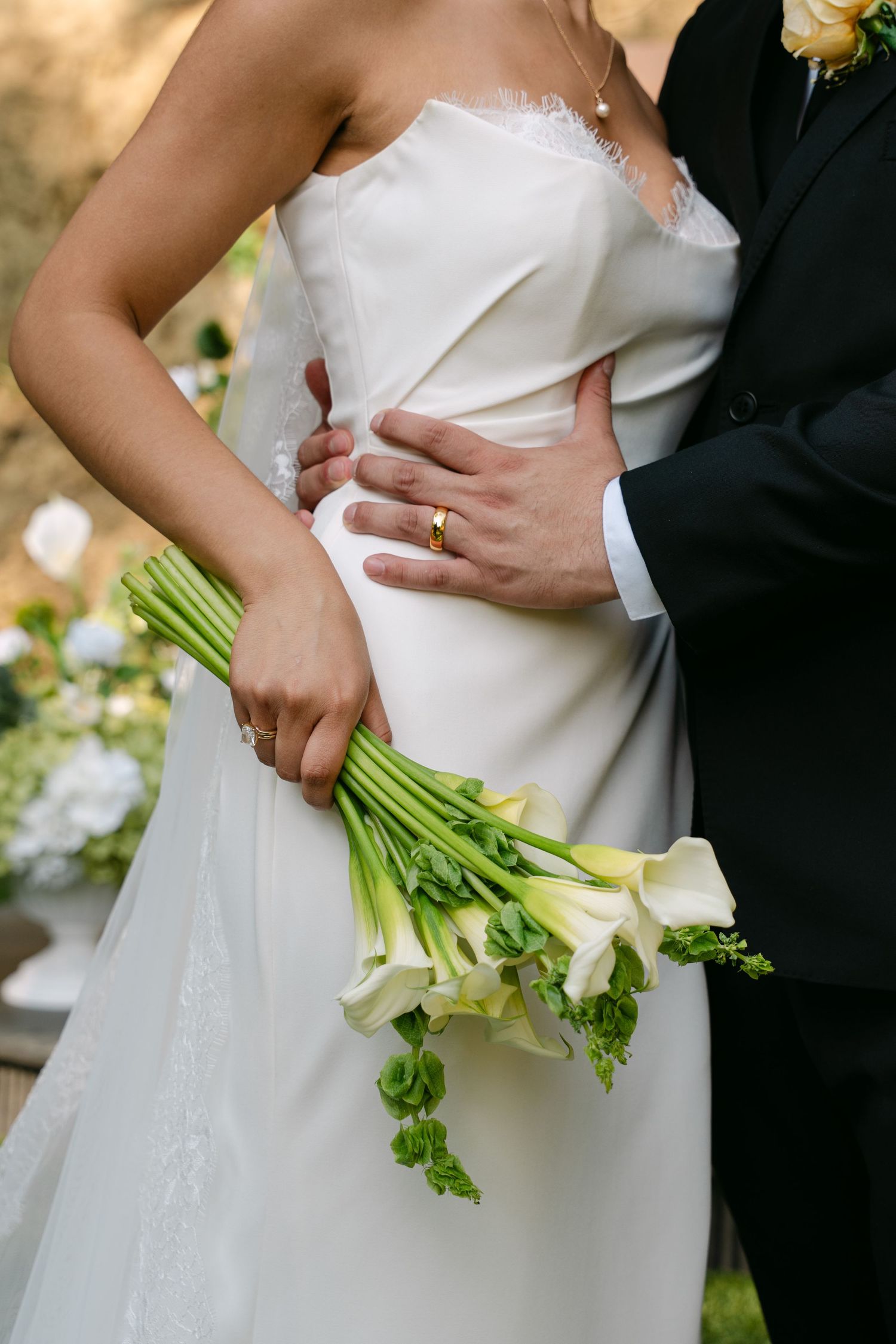 Groom places hand on brides waist as she holds white calla lily bouquet after outdoor ceremony.