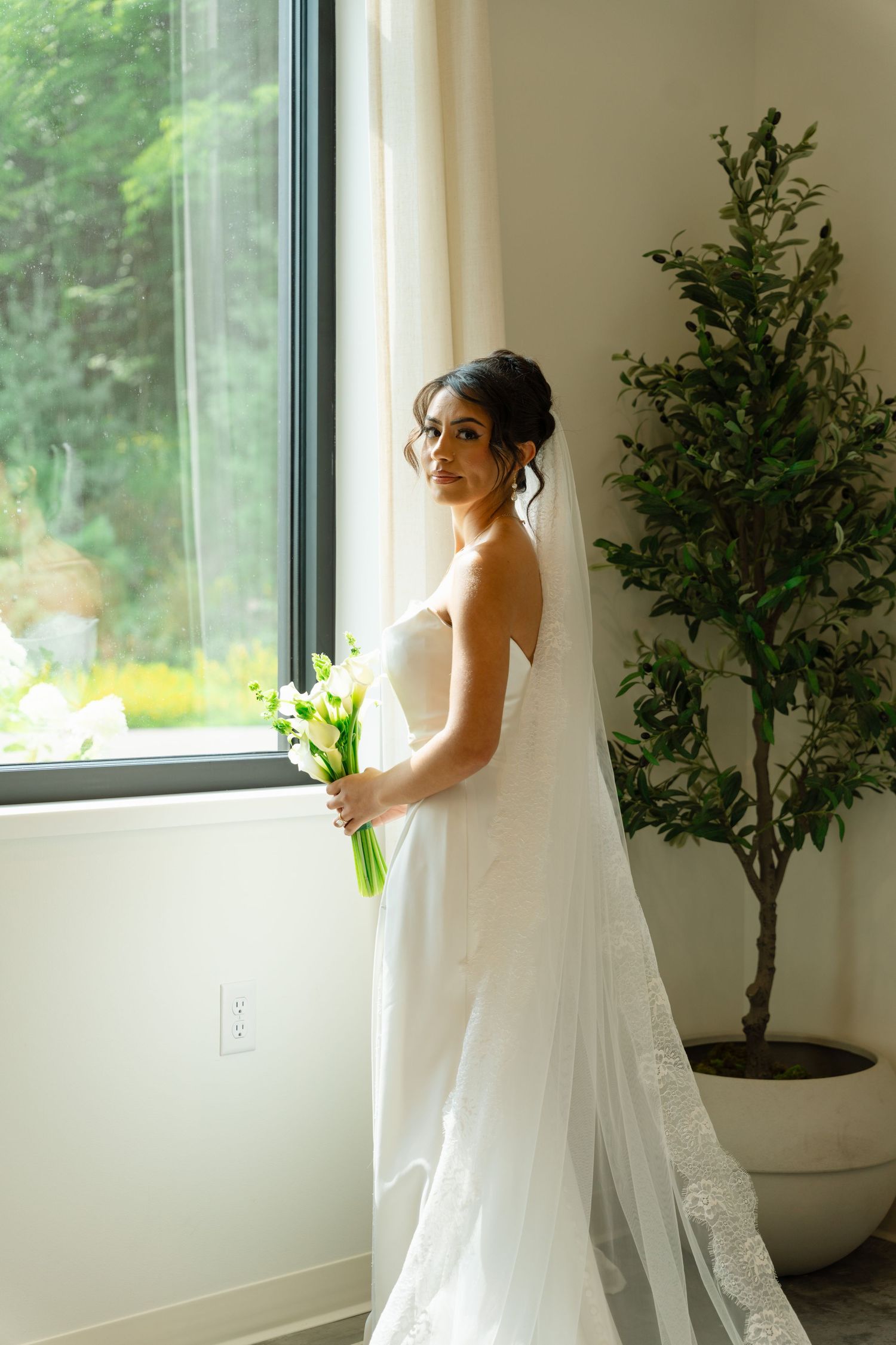 Bride in strapless white gown holding a white calla lily bouquet standing by a large window.