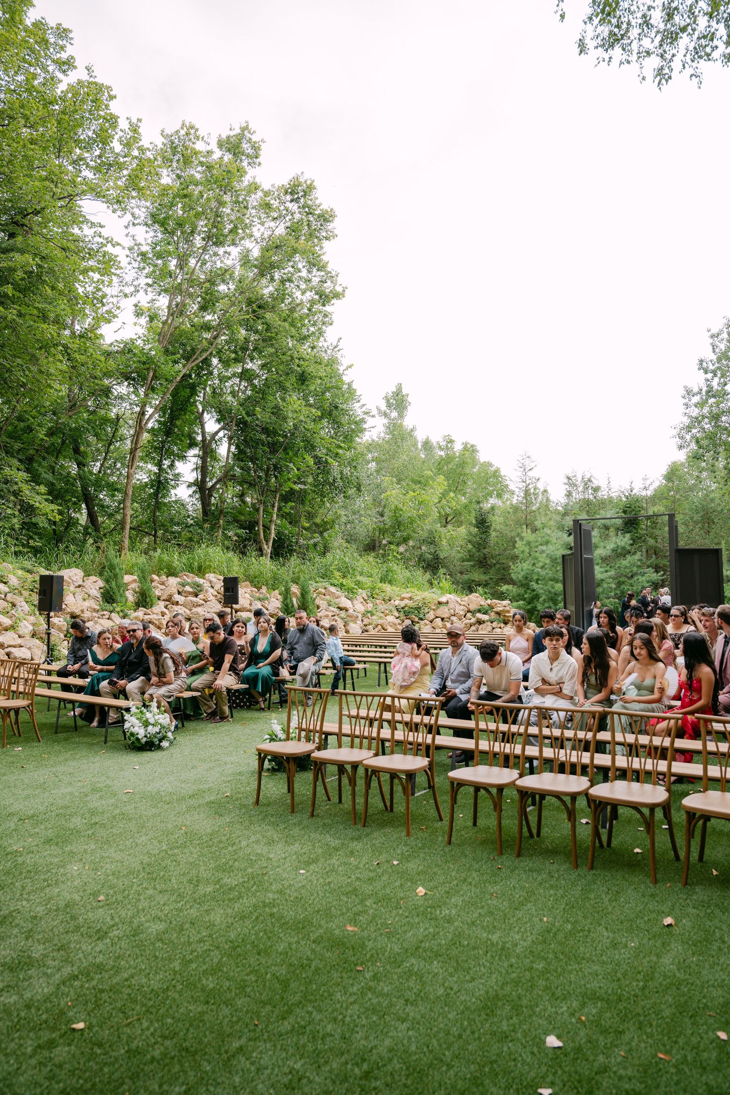 Guests seated on wooden chairs at an elegant outdoor wedding ceremony surrounded by lush green trees.