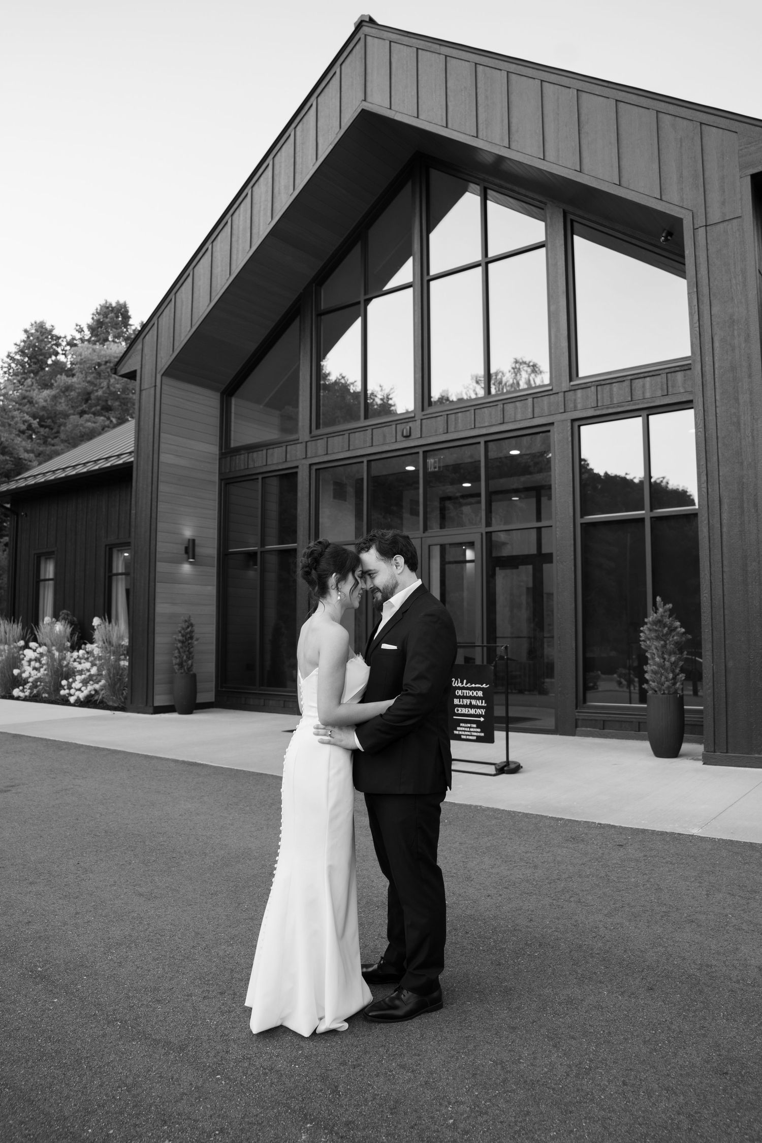 Bride and groom share a romantic moment outside a modern wedding venue in black and white.