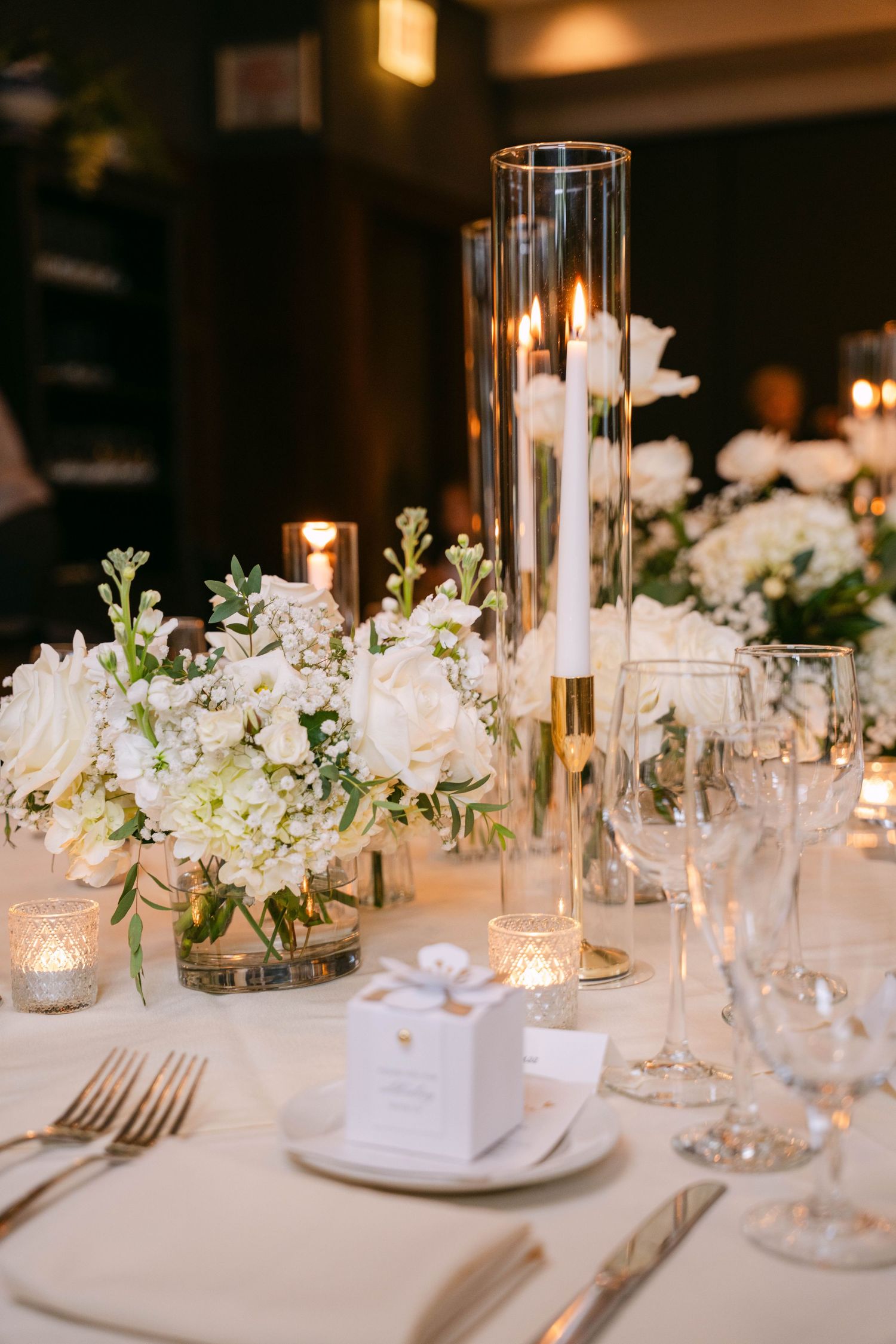 Elegant wedding reception table with white floral centerpieces, tall candles, and white favor boxes.