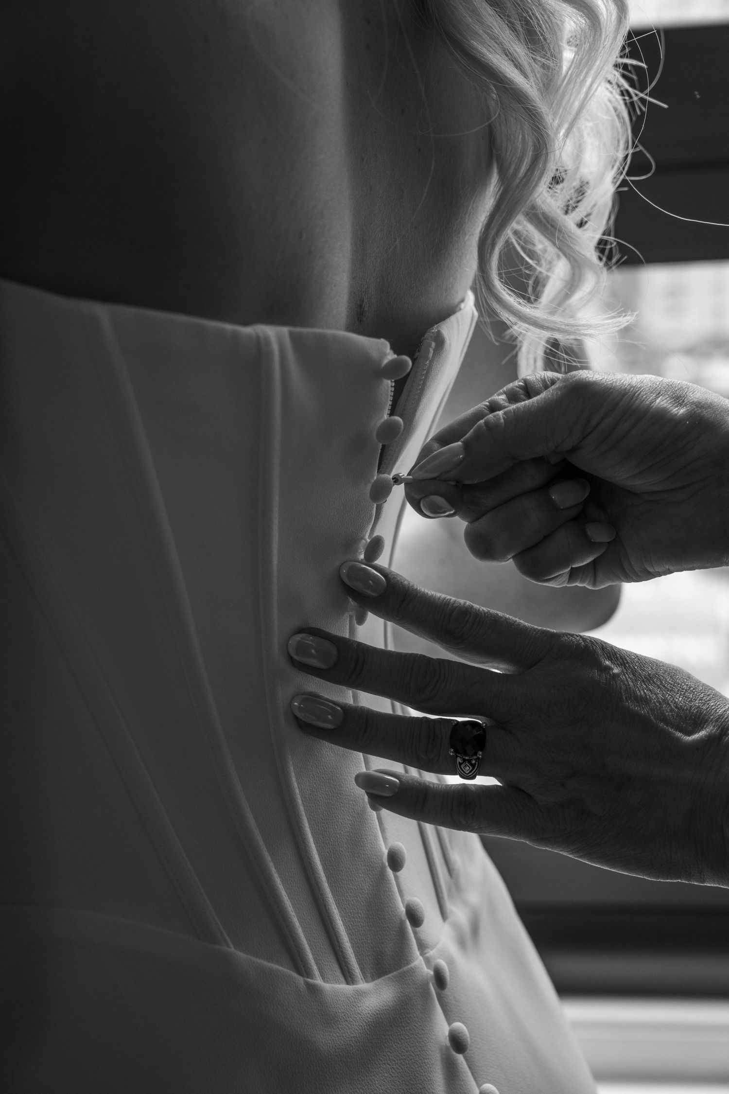 Tattooed hands fastening delicate buttons on back of bride's white wedding dress before ceremony.