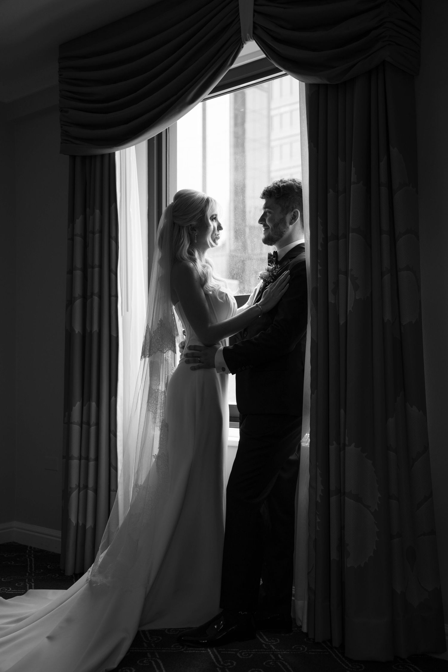 Black and white wedding photo of bride and groom holding hands by draped window, elegant silhouette portrait.