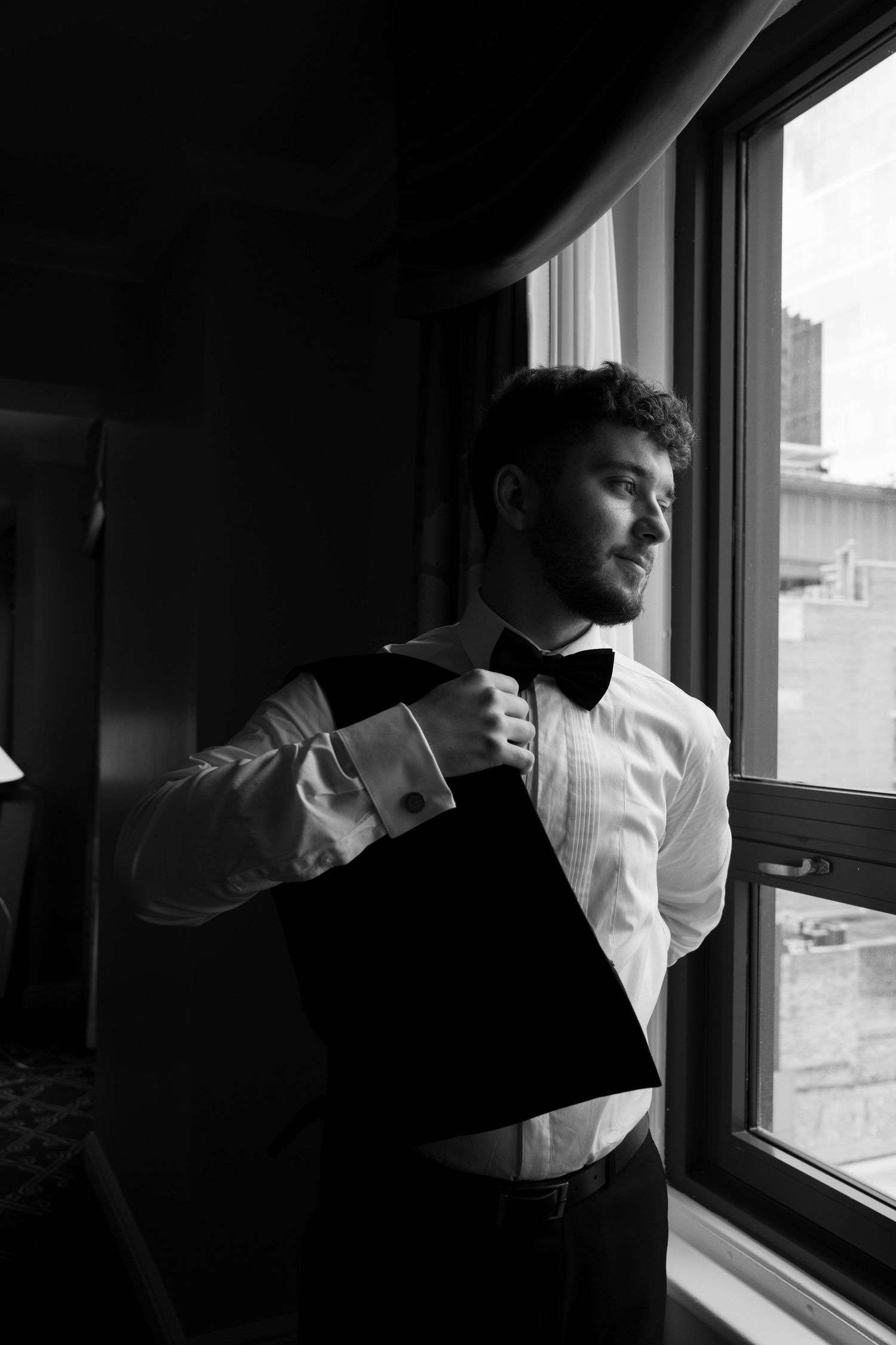Groom in tuxedo shirt and bow tie adjusting his jacket while gazing out a window in black and white.