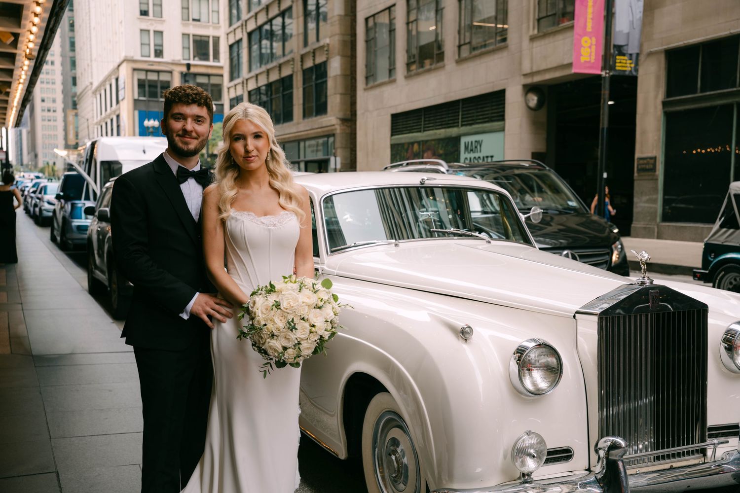 Bride and groom posing beside a classic white Rolls-Royce on a city street, bride holding a white bouquet.