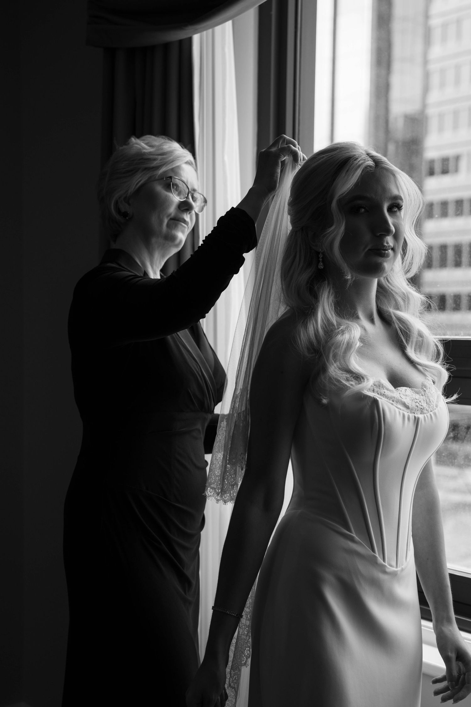 Black and white photo of mother helping bride with veil near window before wedding ceremony.