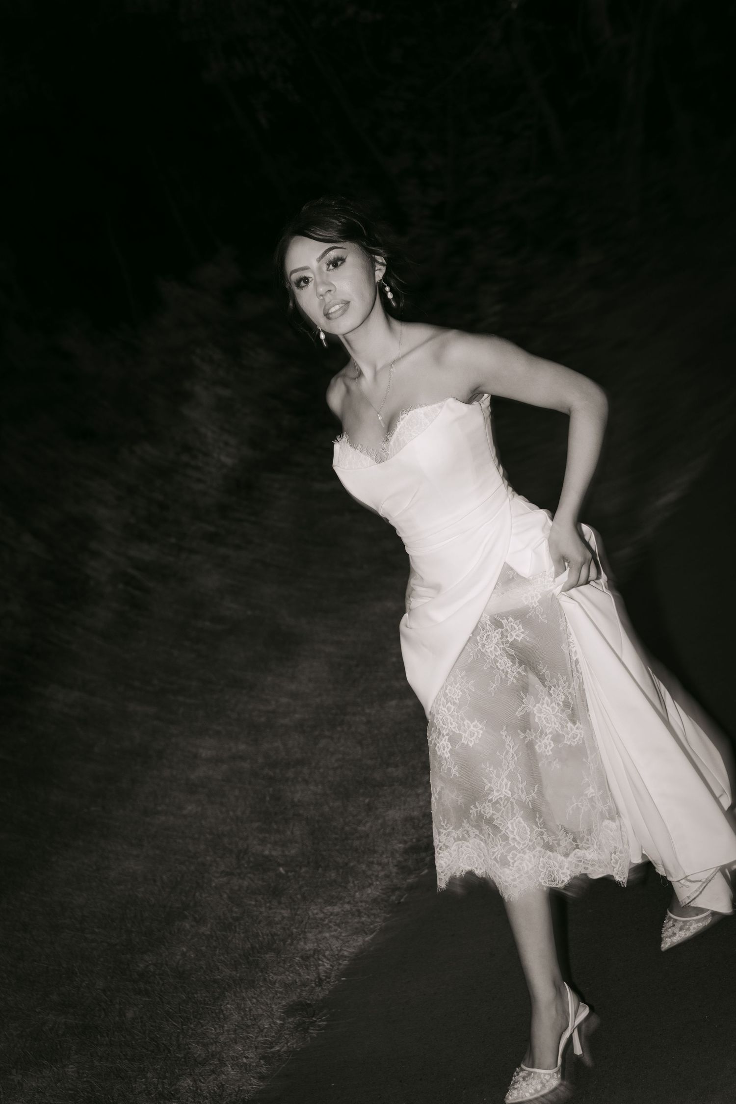 Black and white photo of bride in strapless lace wedding gown walking along dark outdoor path.
