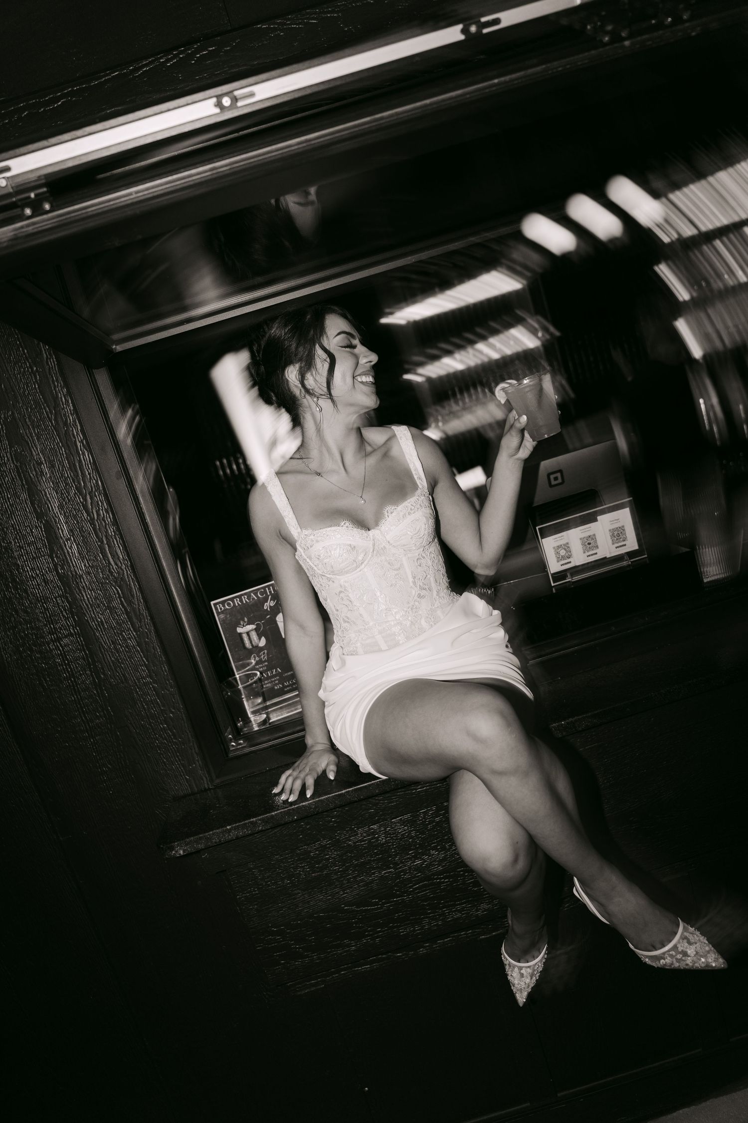 Joyful bride in short white dress laughing while seated at a bar during wedding reception.