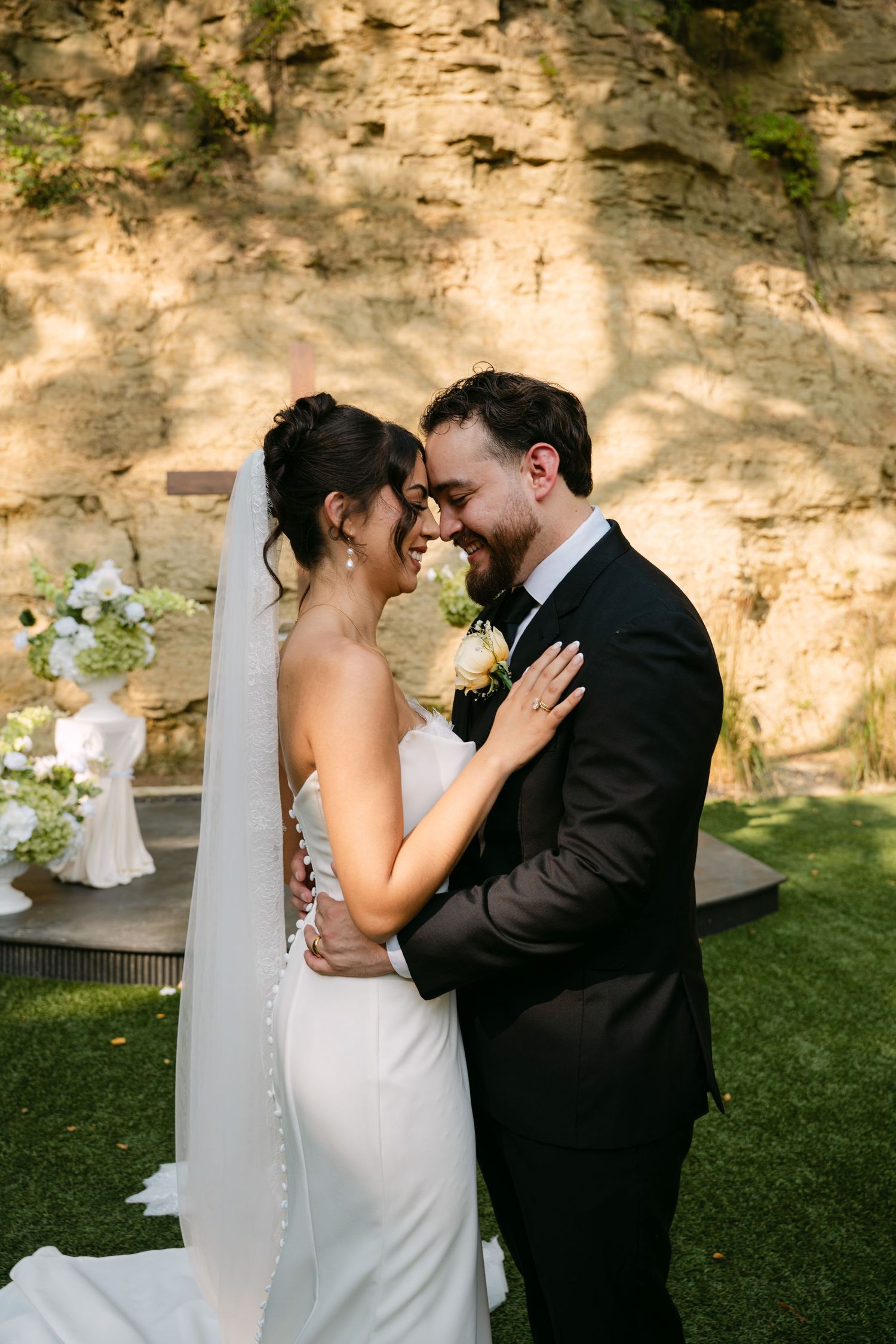 Bride and groom sharing an intimate forehead-to-forehead moment in front of a sunlit stone wall.