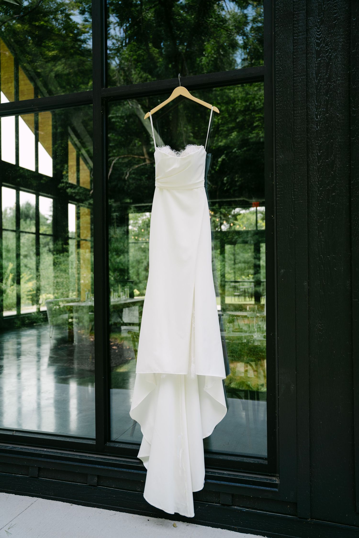Elegant white wedding dress hanging on a wooden hanger against dark glass doors with garden view.
