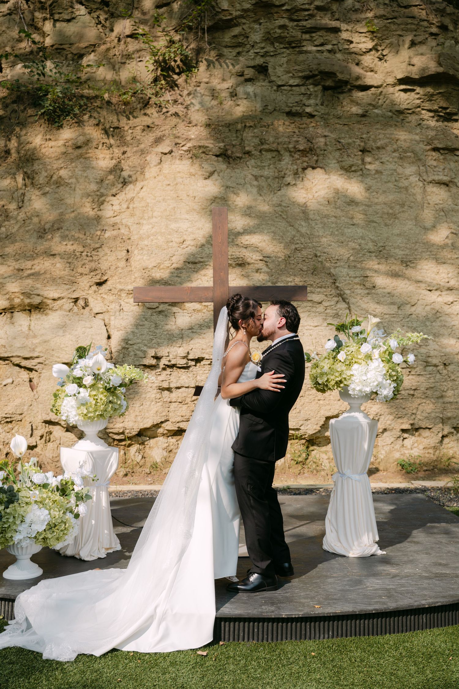 Newlyweds share romantic first kiss at outdoor altar with cross and white hydrangea arrangements.