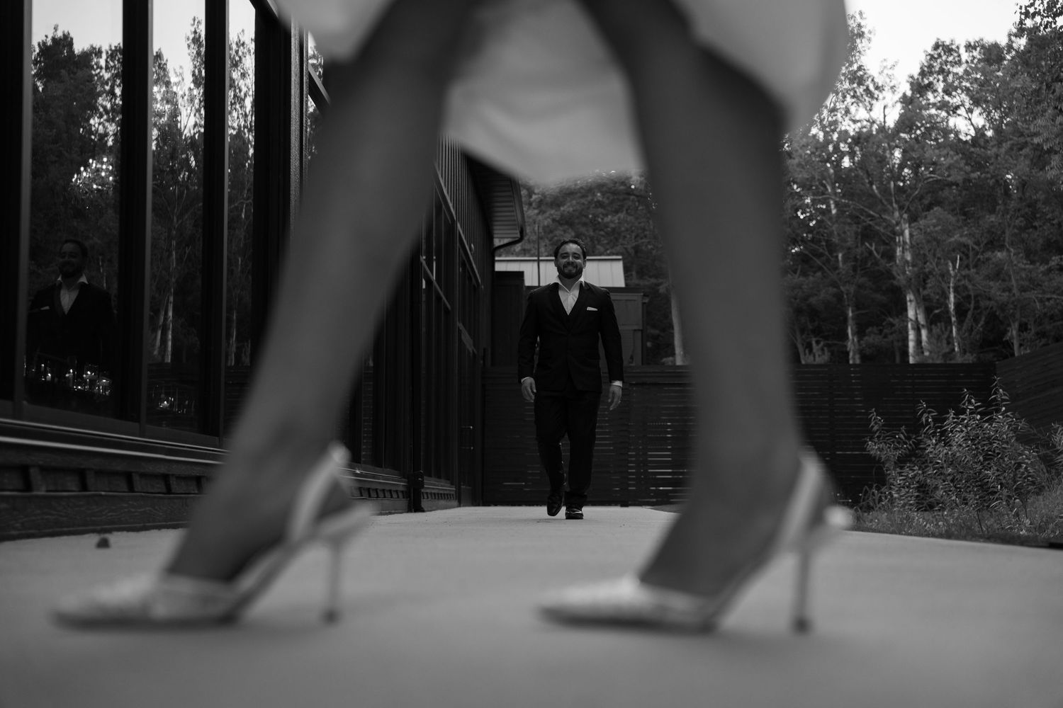Artistic black and white wedding photo of groom approaching bride, framed by her heels in foreground.