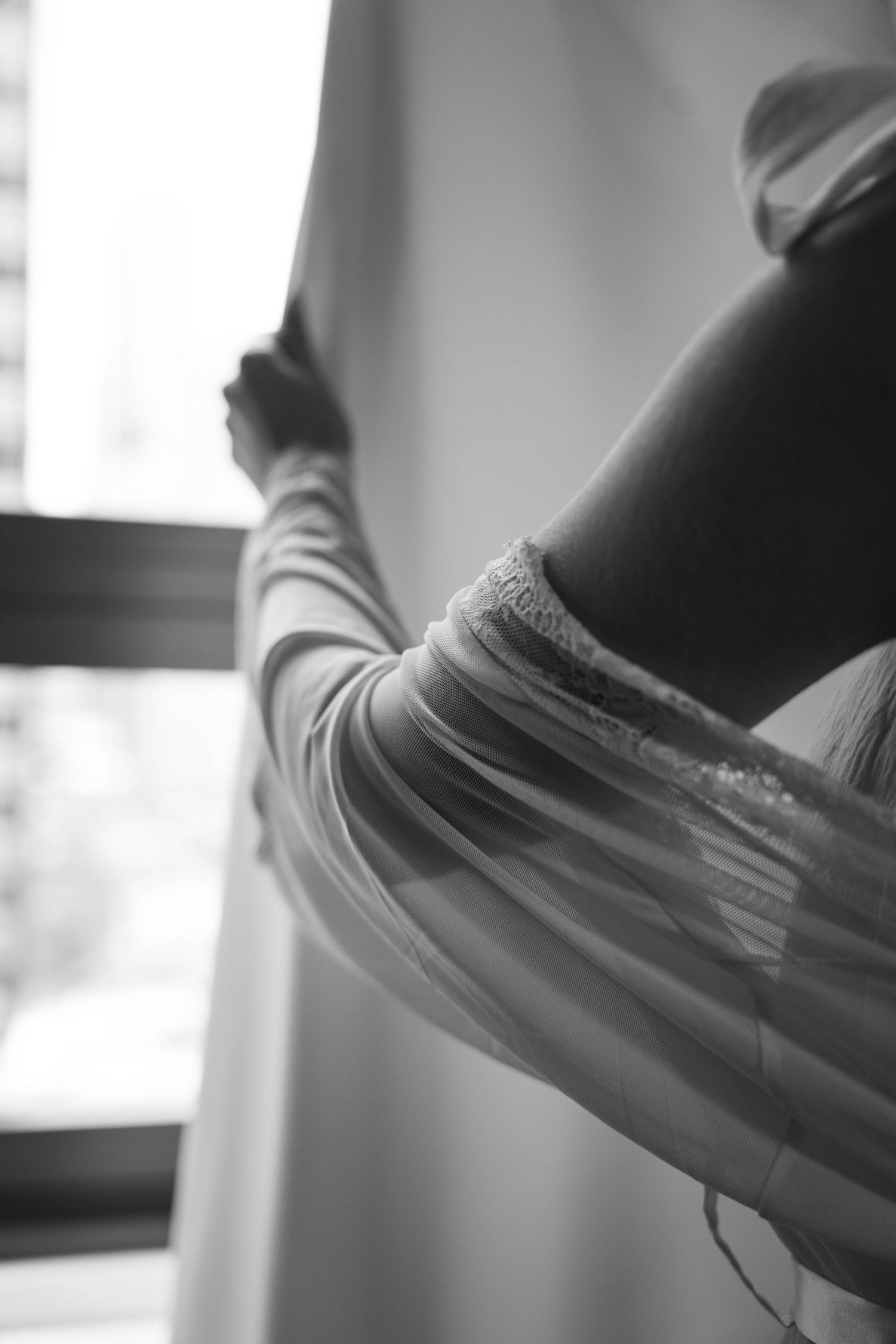 Black and white photo of bride in lace robe holding curtain near window.