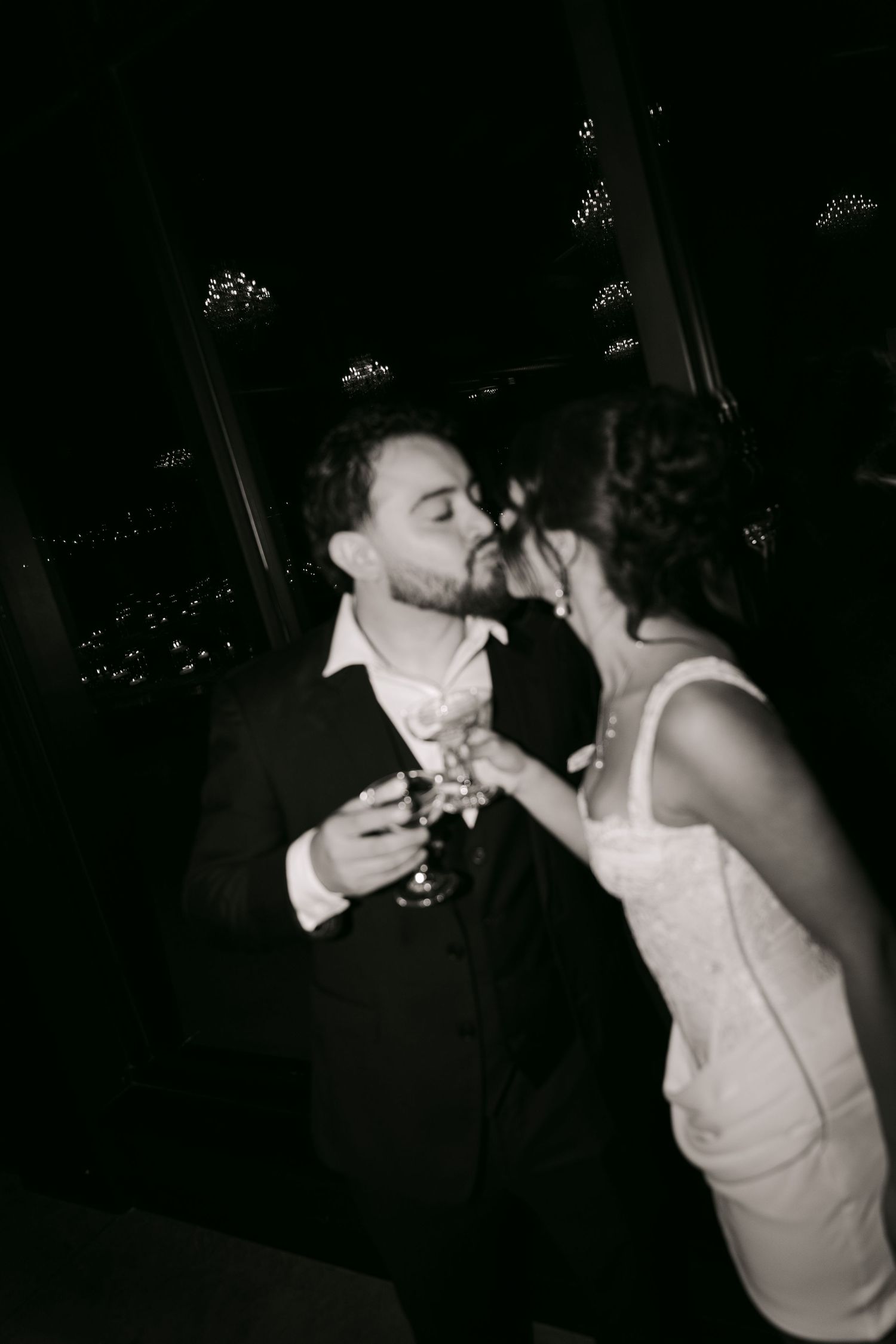 Black and white photo of bride and groom sharing a kiss while holding champagne at wedding.
