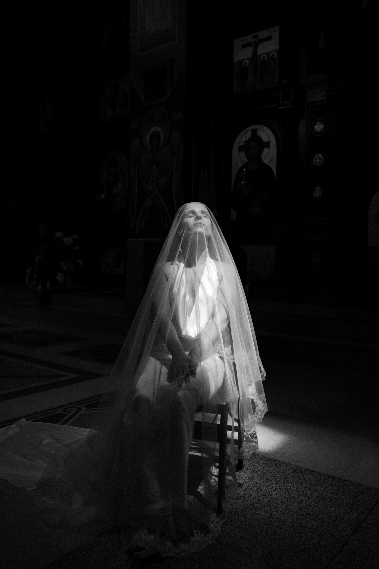 Black and white photo of a bride in veil sitting in a church, illuminated by a beam of light.