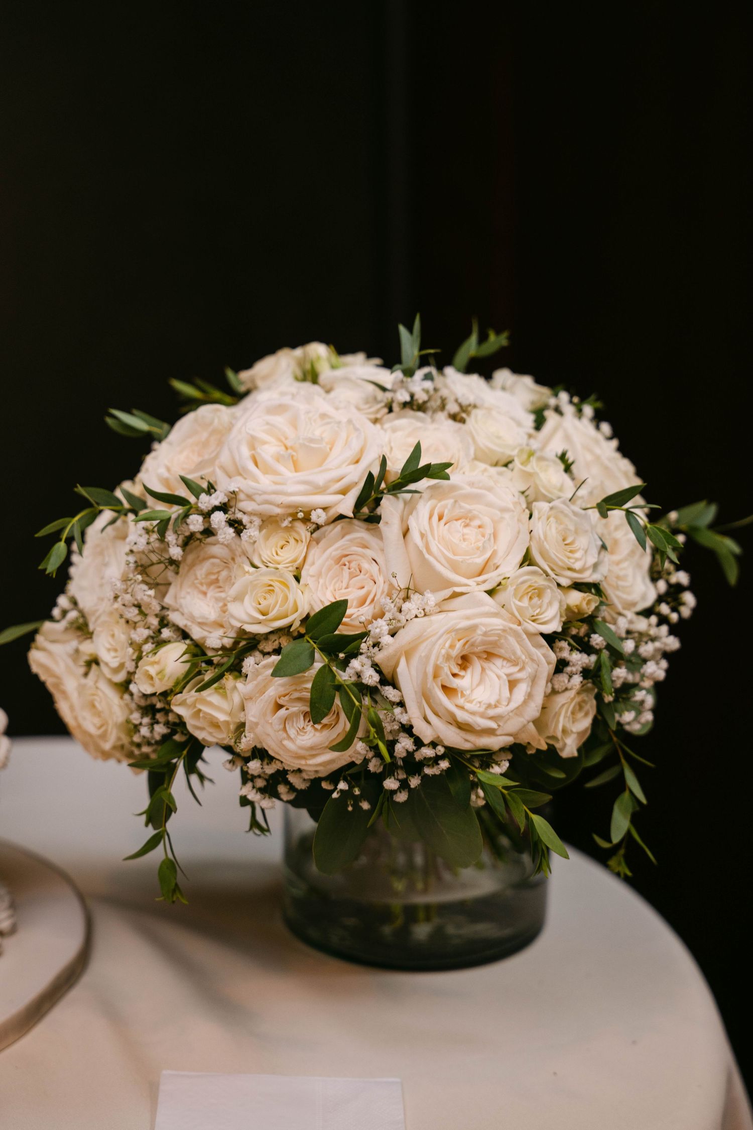 Elegant white rose centerpiece with baby's breath and greenery in a glass vase on a white table.