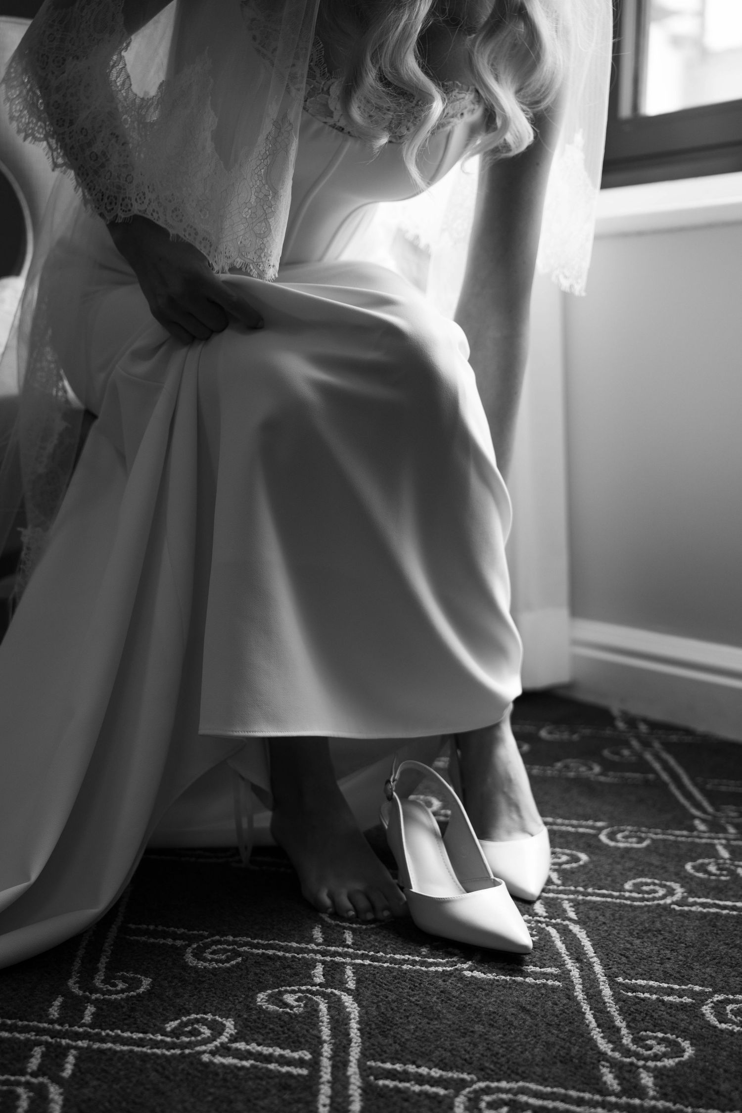 Bride in white wedding gown sitting by window with white heels on patterned carpet, black and white.