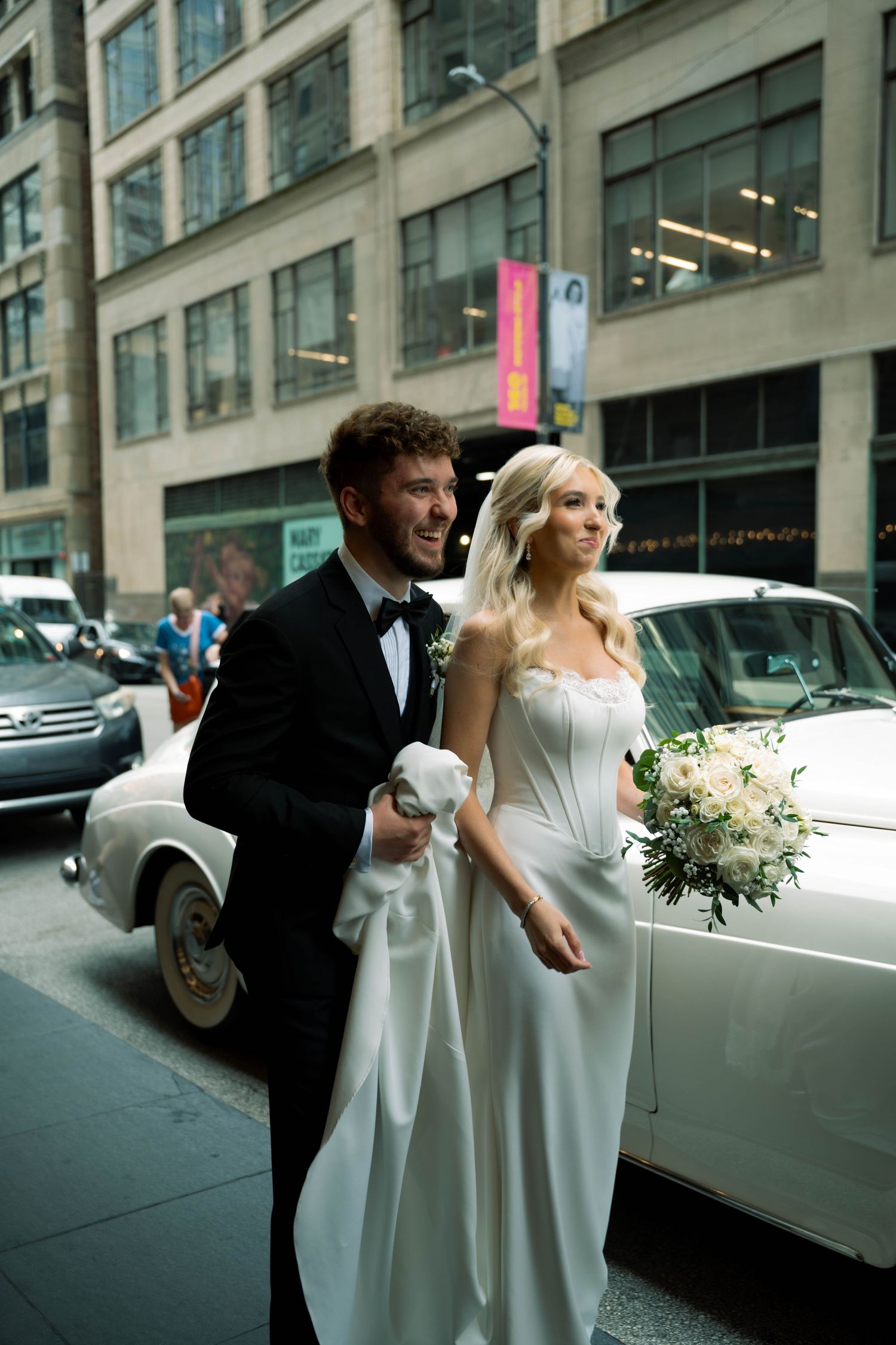 Smiling bride and groom in wedding attire walking together on a city street beside a vintage white car.