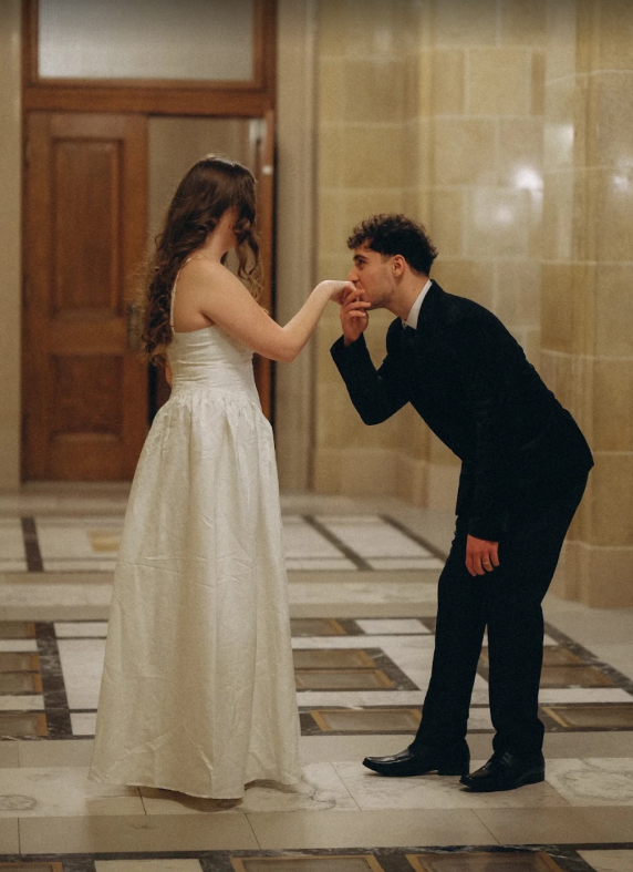 Groom in black suit kissing bride's hand in elegant marble-floored hallway.