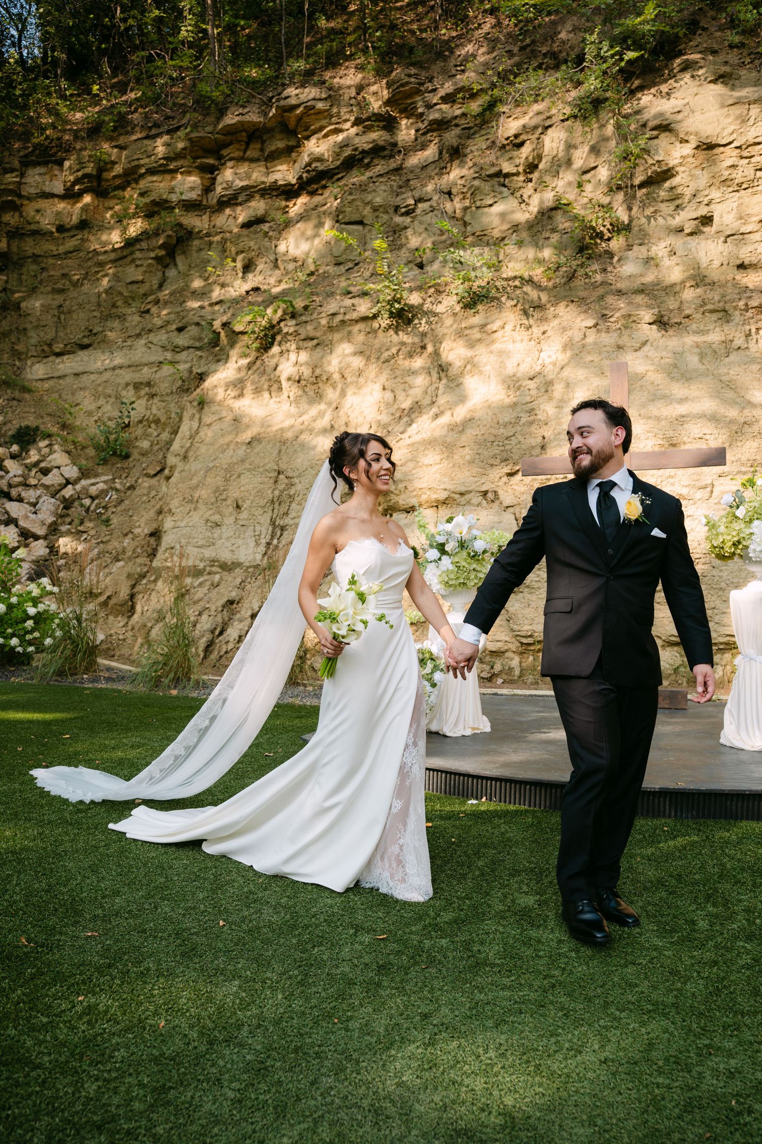 Smiling newlyweds holding hands walking away from outdoor wedding altar with stone cliff backdrop.