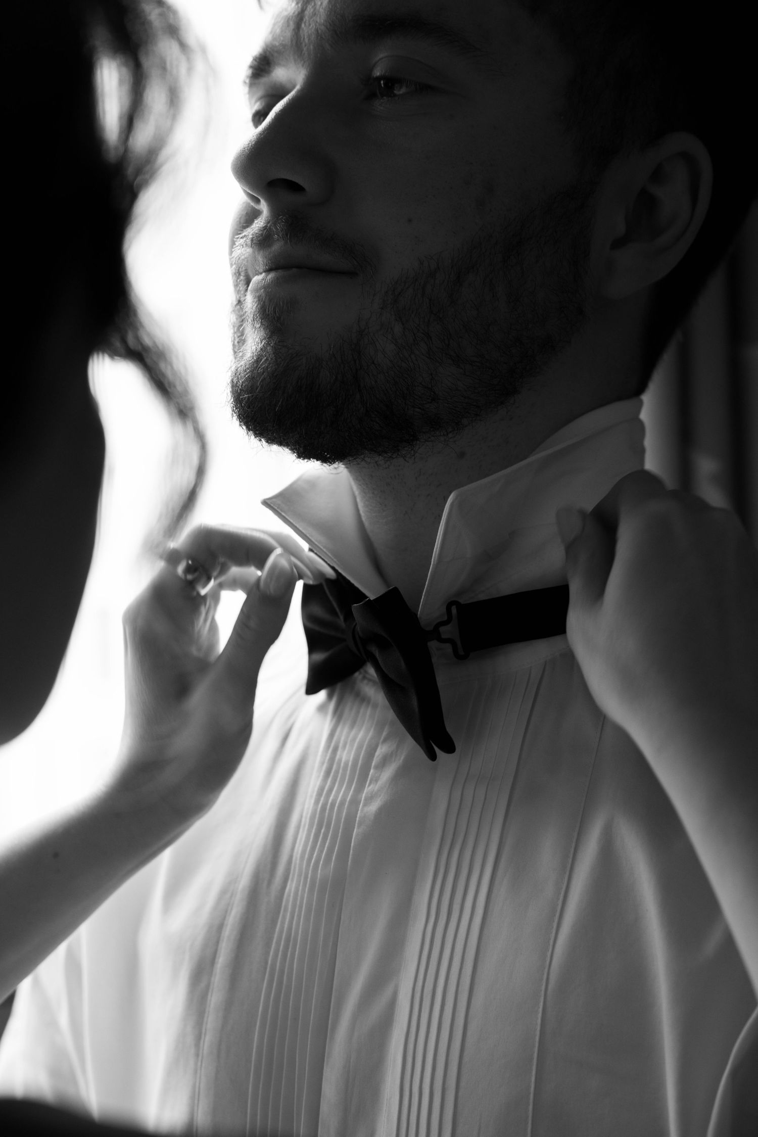 Groom in white tuxedo shirt having black bow tie adjusted by hands before wedding ceremony.