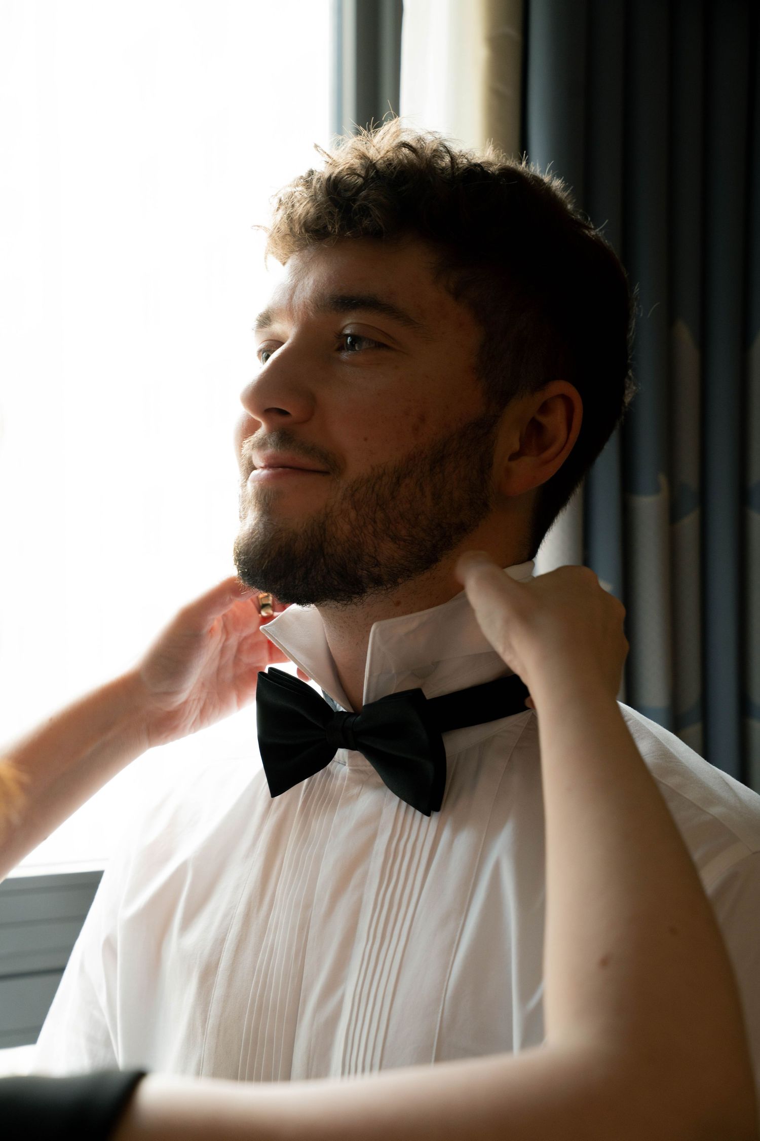 Smiling groom in white tuxedo shirt and black bow tie being dressed by hands adjusting his collar near a bright window.