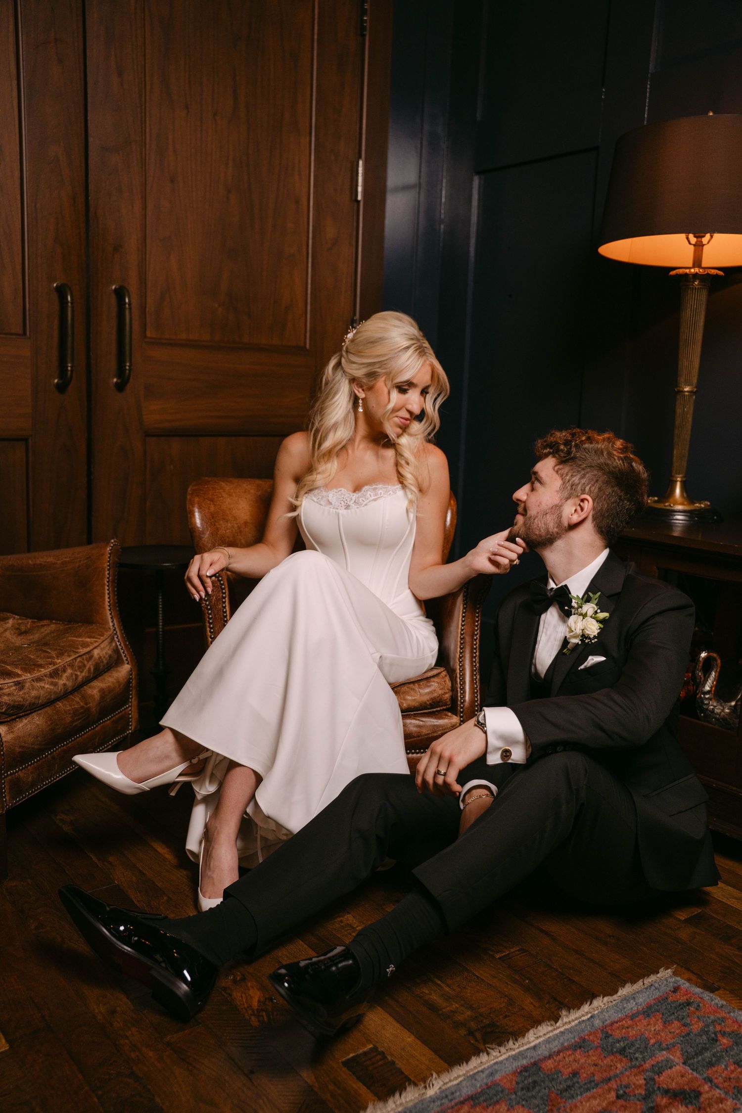 Bride seated in leather chair with groom at her feet in warmly lit room.