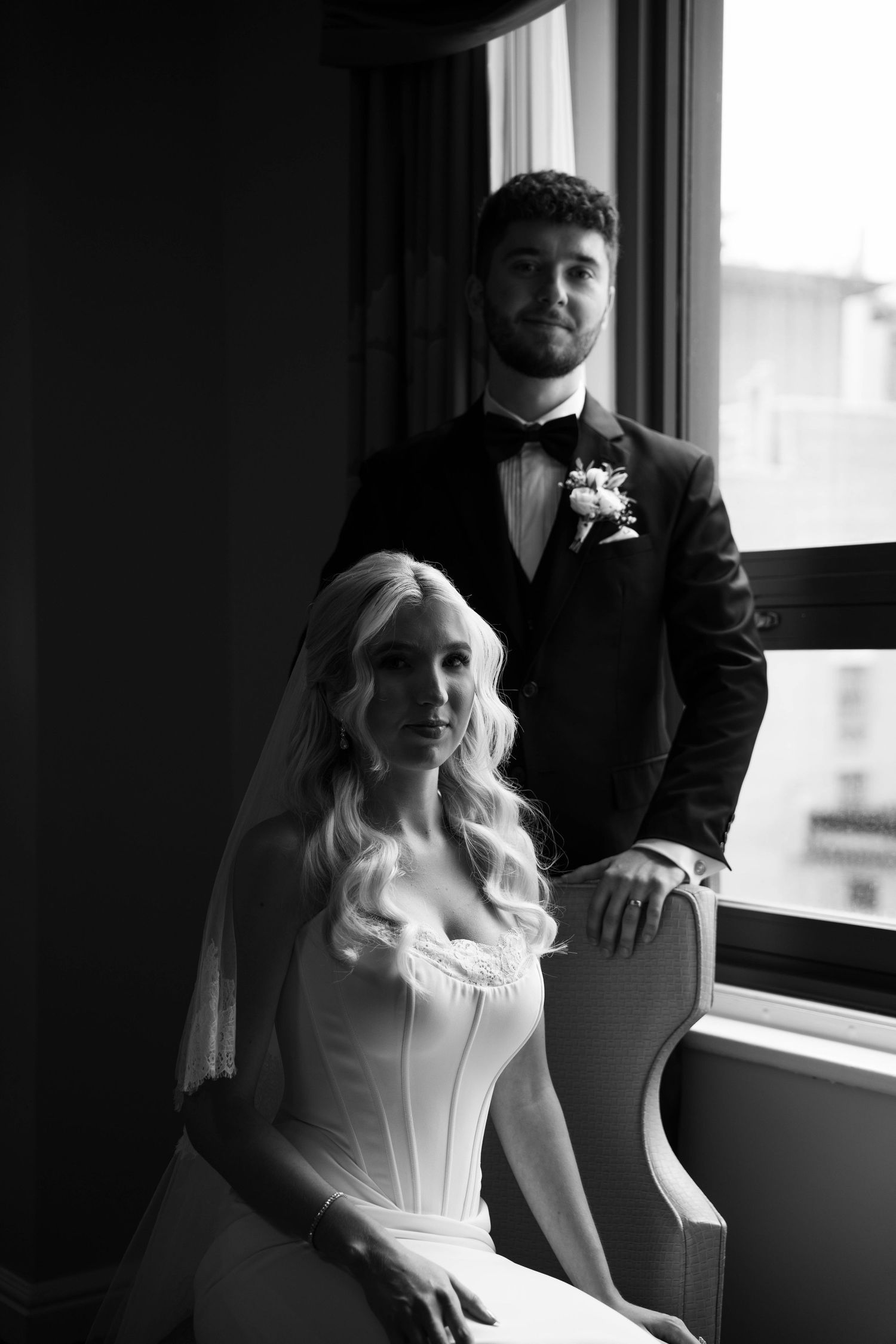 Black and white portrait of bride and groom posing by window in hotel room.
