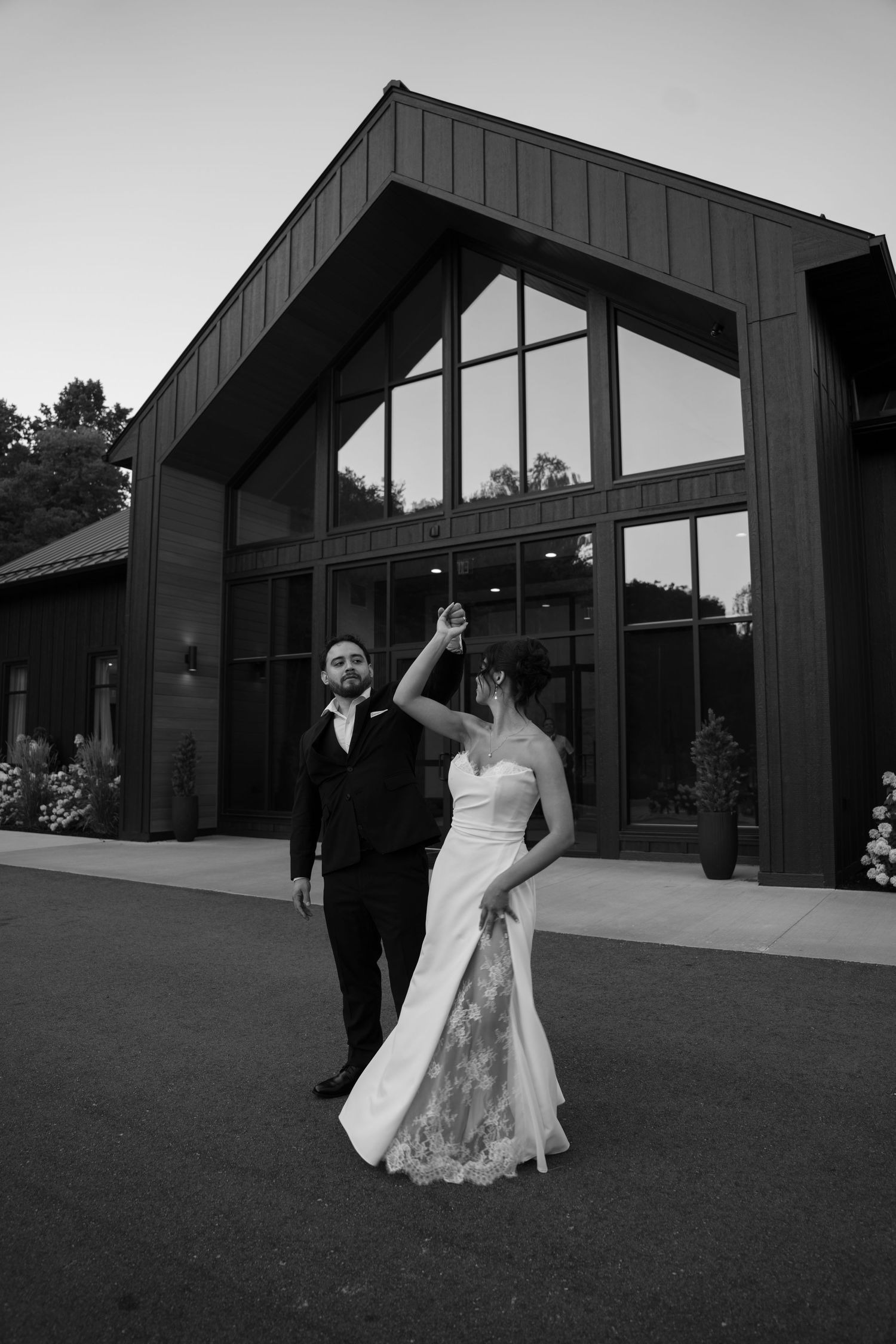 Groom spins bride in a joyful dance outside a modern wedding venue at twilight.
