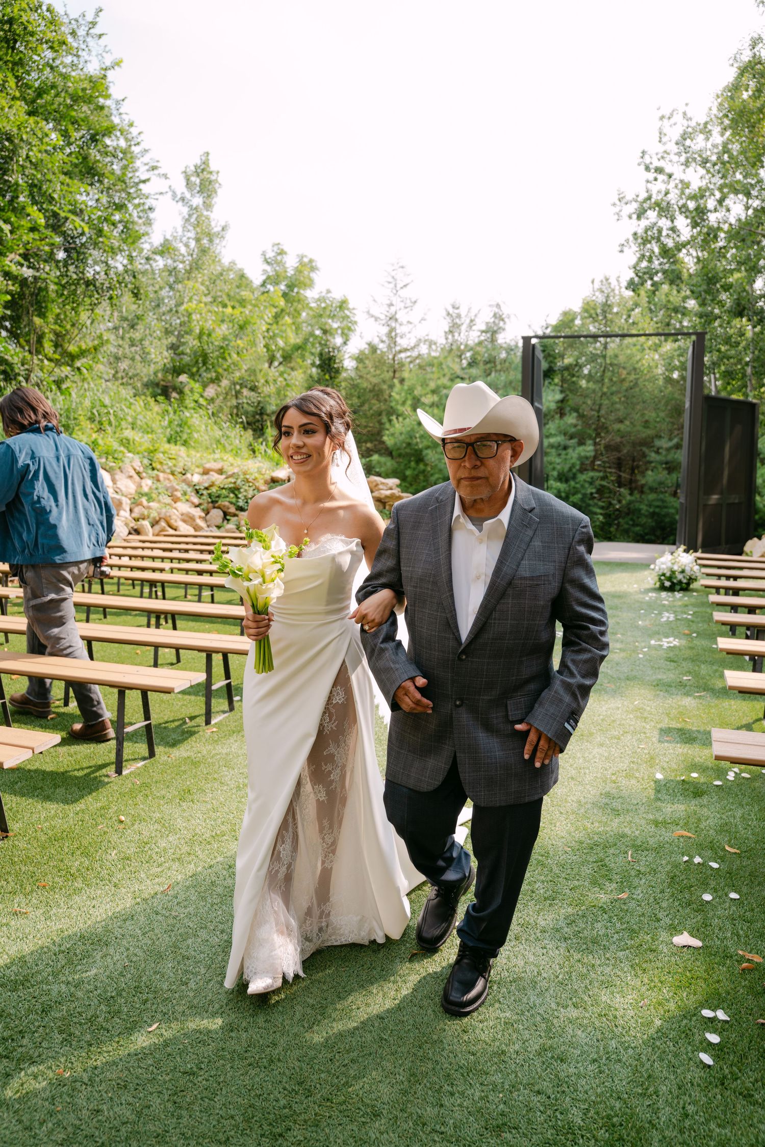 Bride in elegant white gown with lace detail walks down outdoor aisle with father in cowboy hat.