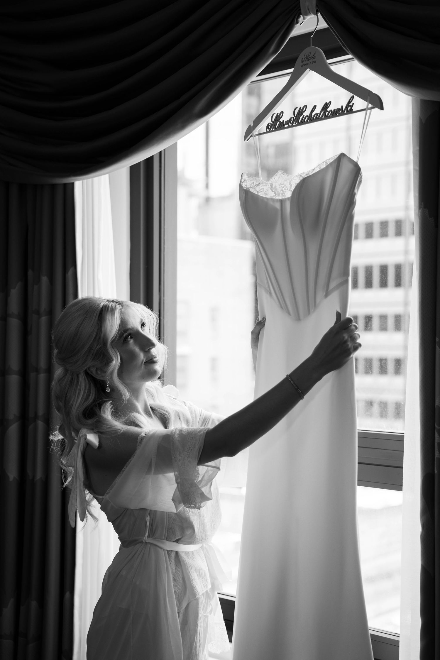 Blonde bride in robe admiring her wedding gown hanging on personalized hanger by hotel window.