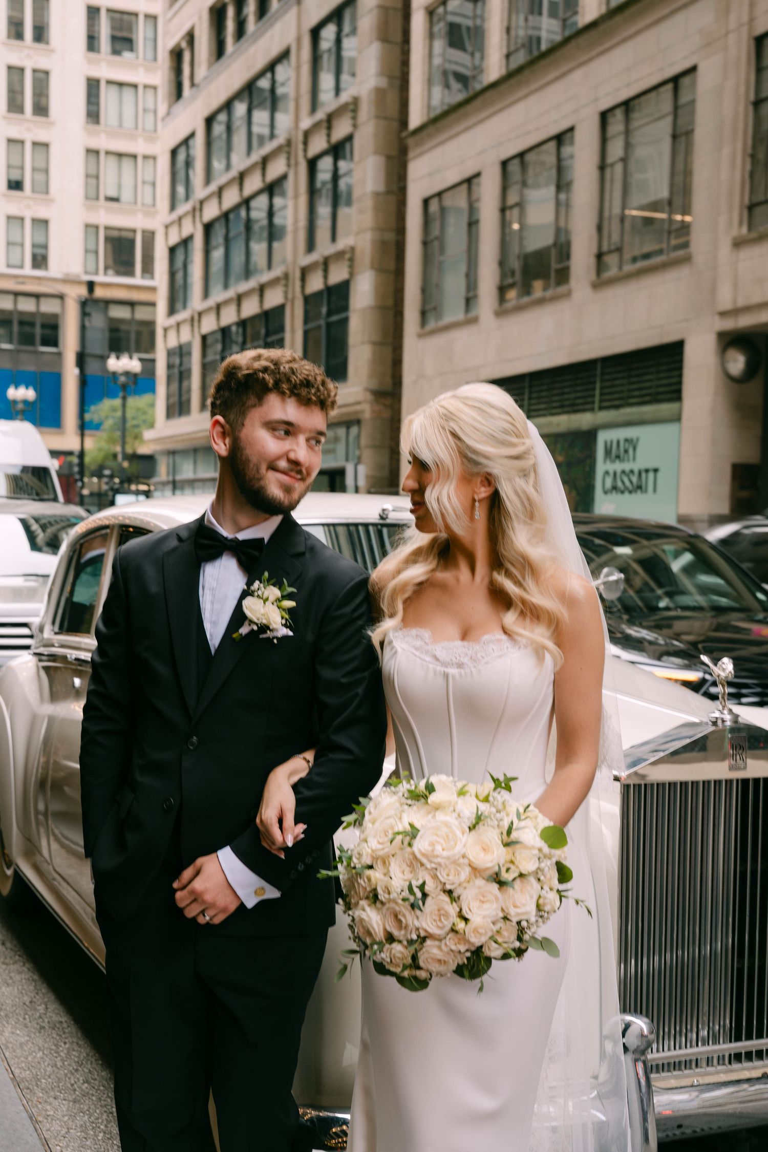 Bride holding white rose bouquet and groom in black tuxedo posing beside a vintage Rolls-Royce on city street.