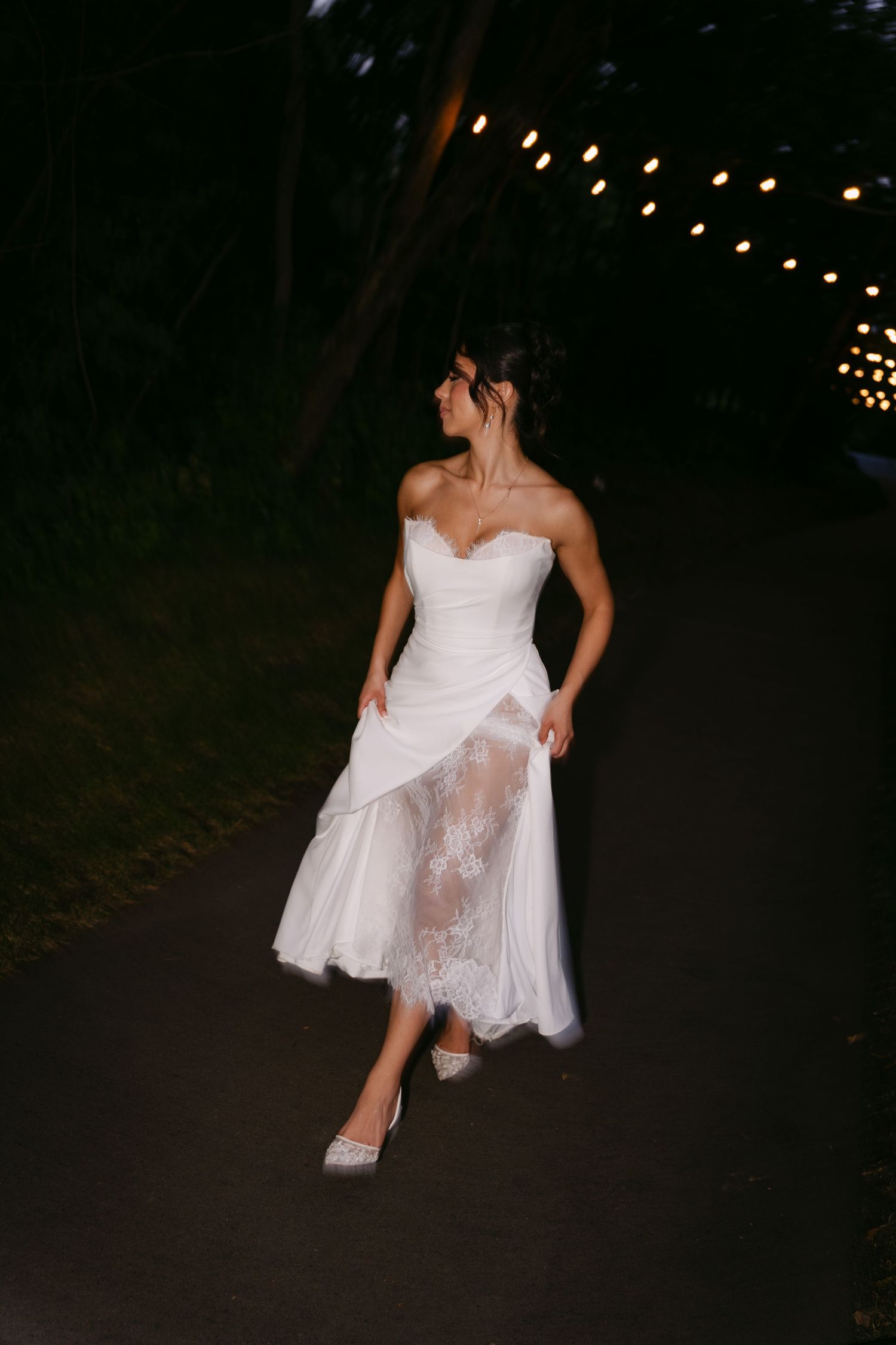 Bride in strapless white lace wedding dress walks along dark path under string lights at night.