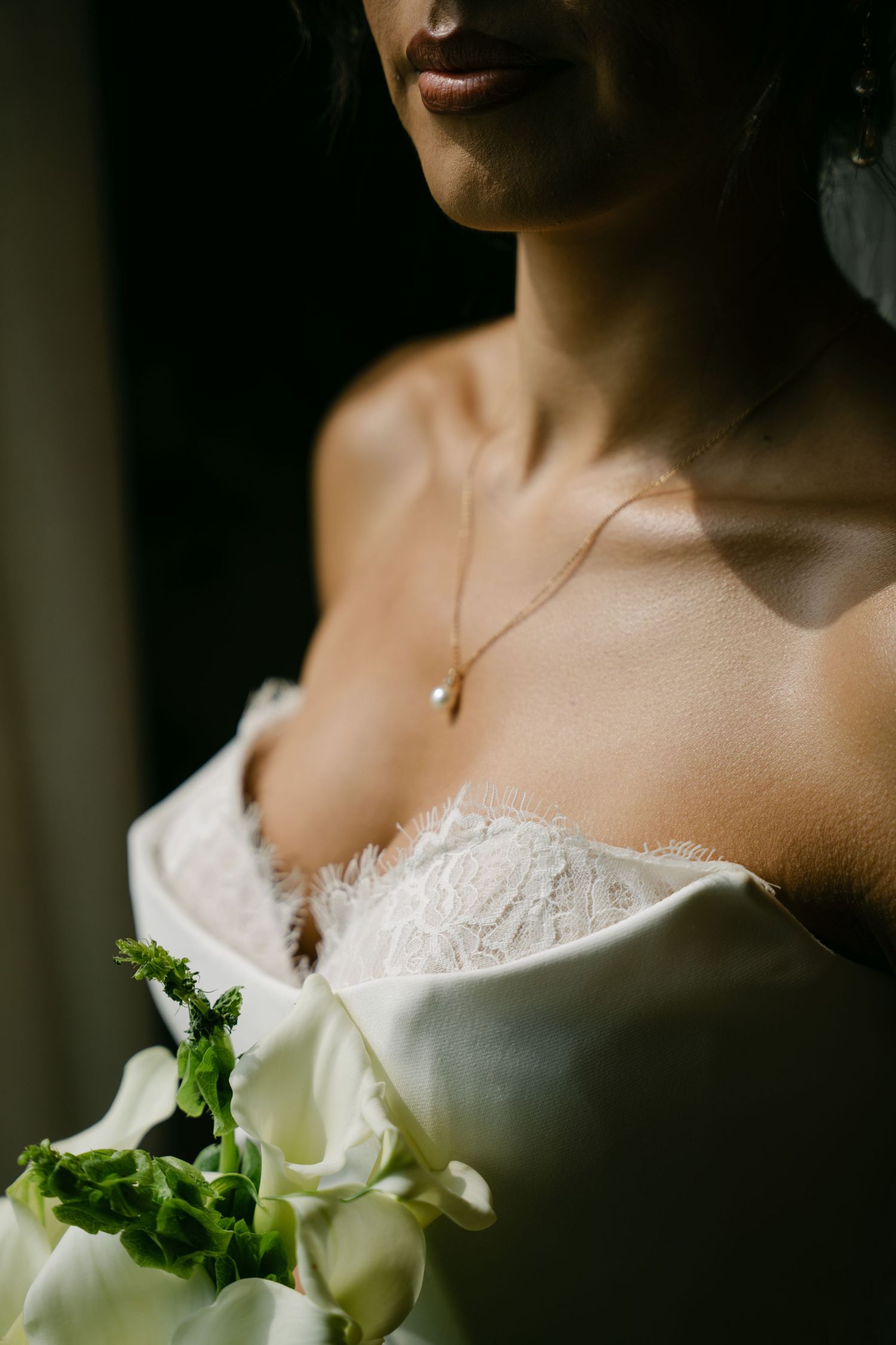 Close-up of bride in strapless lace wedding dress wearing a pearl drop necklace holding bouquet.