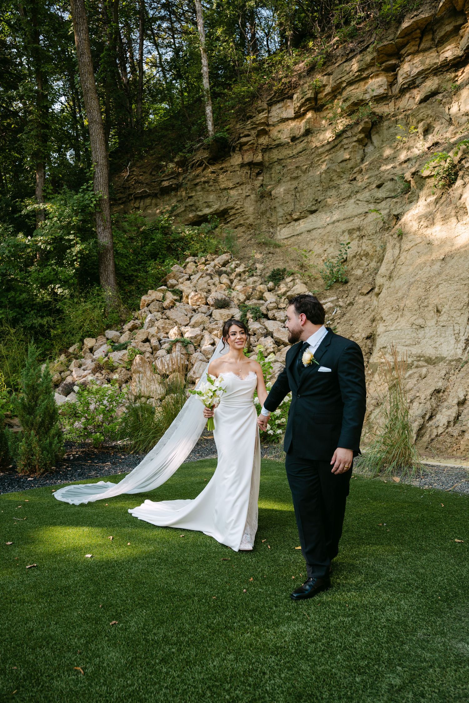 Bride in white gown with veil and groom in black tuxedo walking hand in hand on grass near stone cliff.
