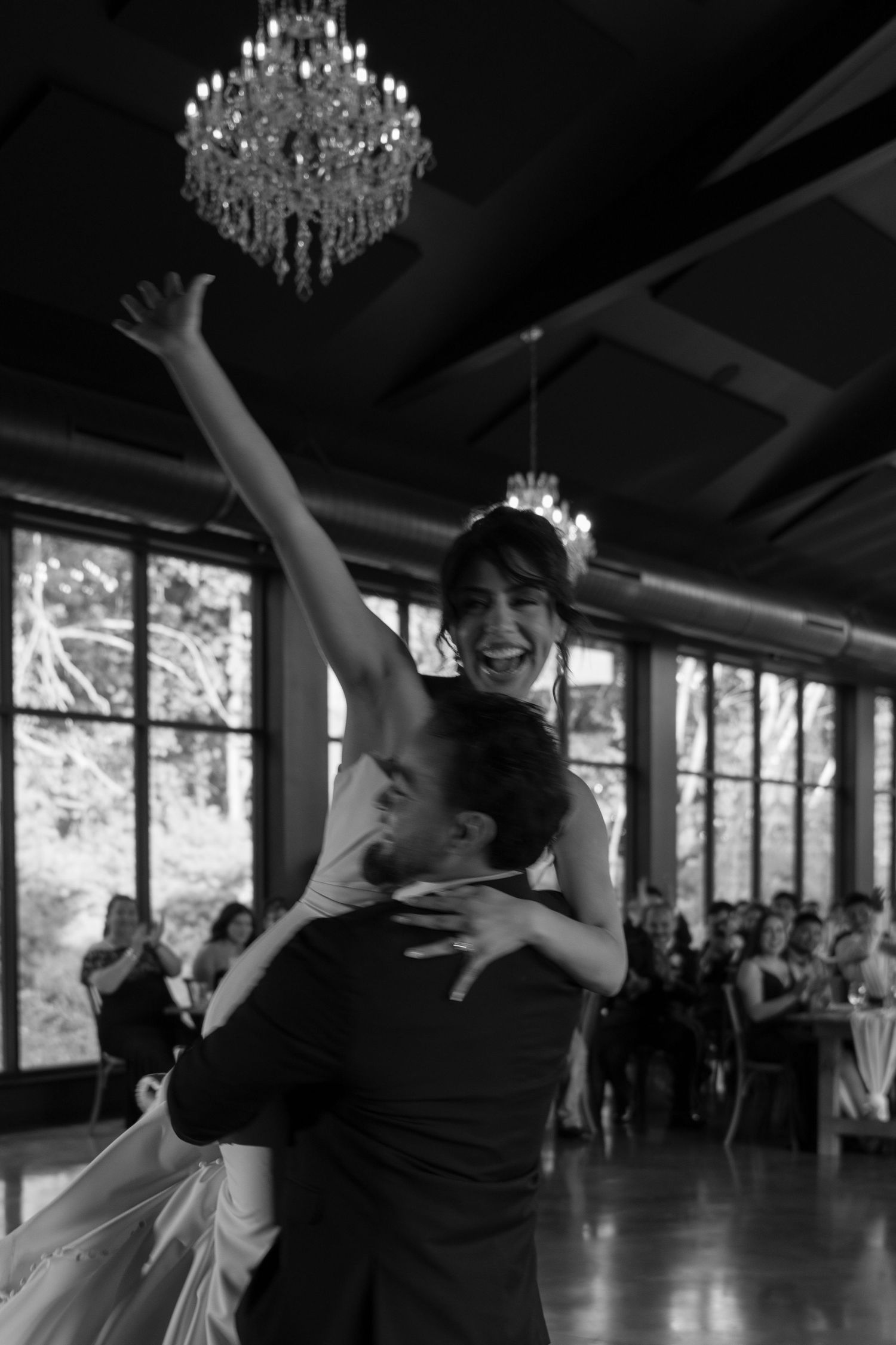 Joyful bride is lifted by groom during first dance under chandelier in black and white wedding reception photo.