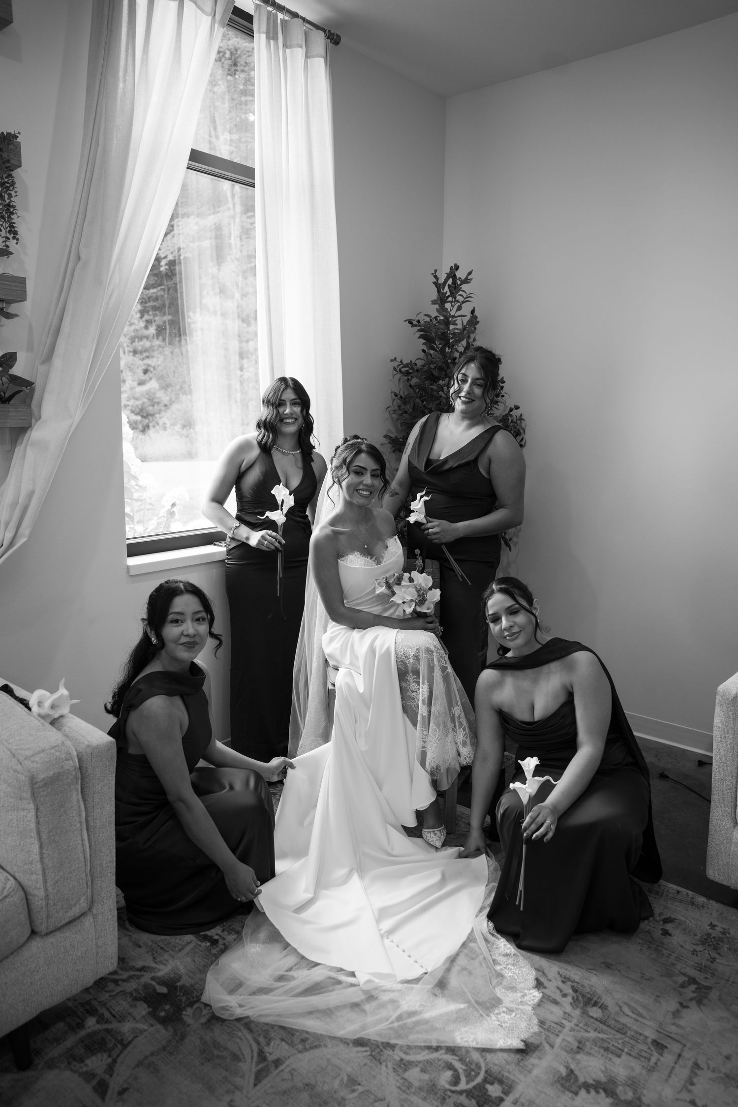 Black and white photo of bride in white gown with four bridesmaids in dark dresses posing by window.