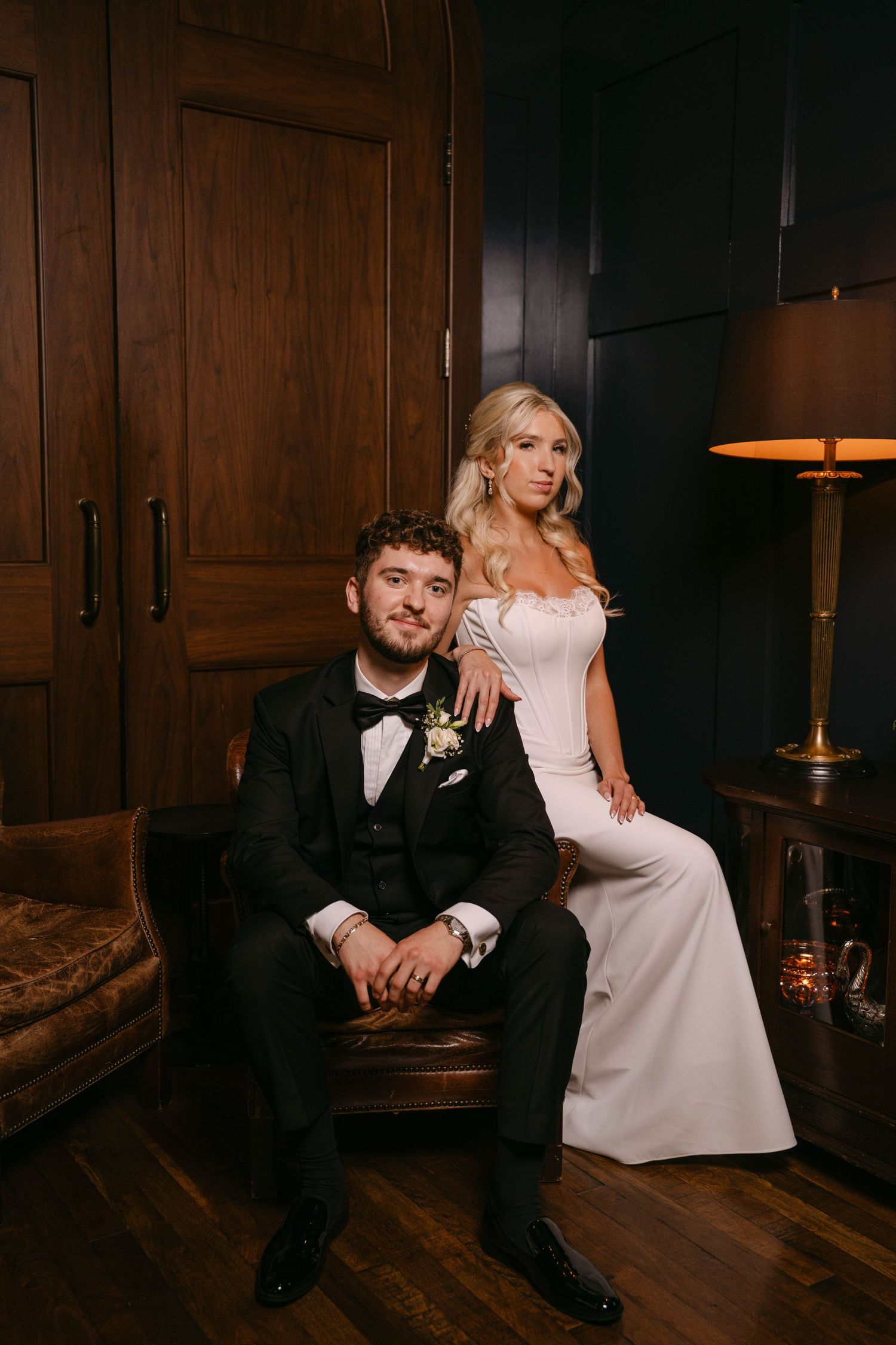Bride in white gown and groom in black tuxedo pose together in a moody dark interior setting.