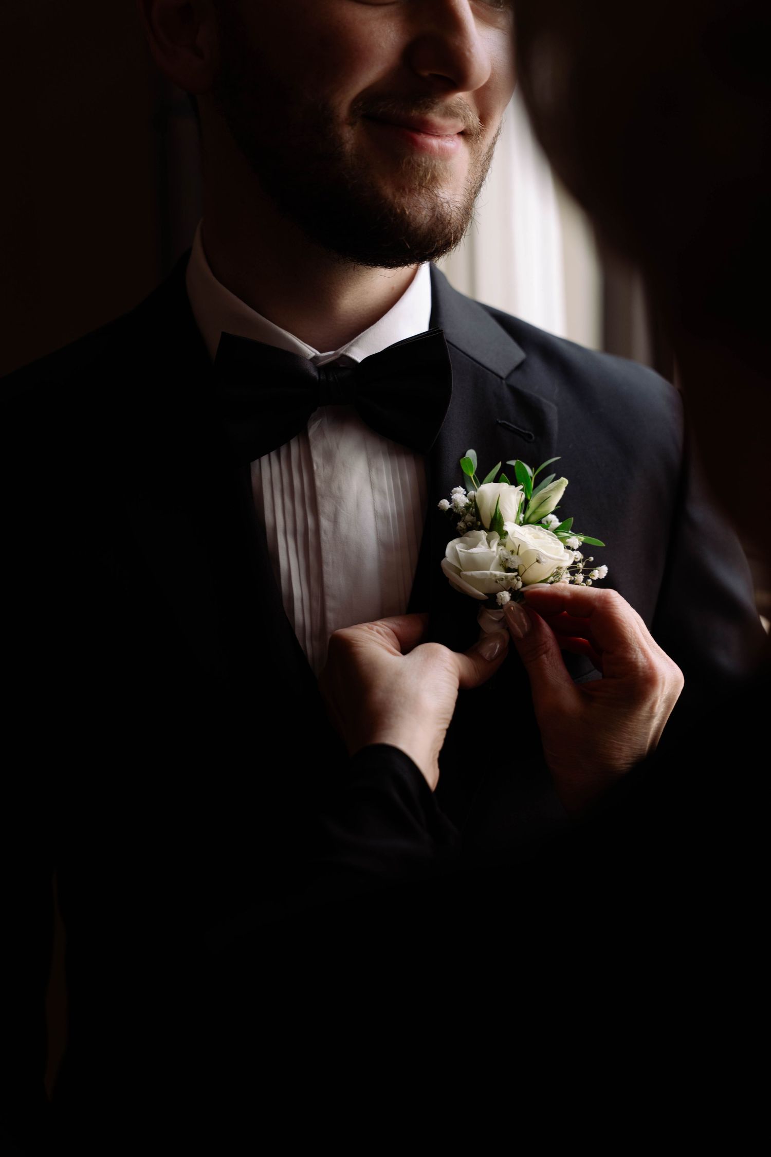 Hands pin a white floral boutonniere on a smiling groom in a black tuxedo in this wedding detail shot.
