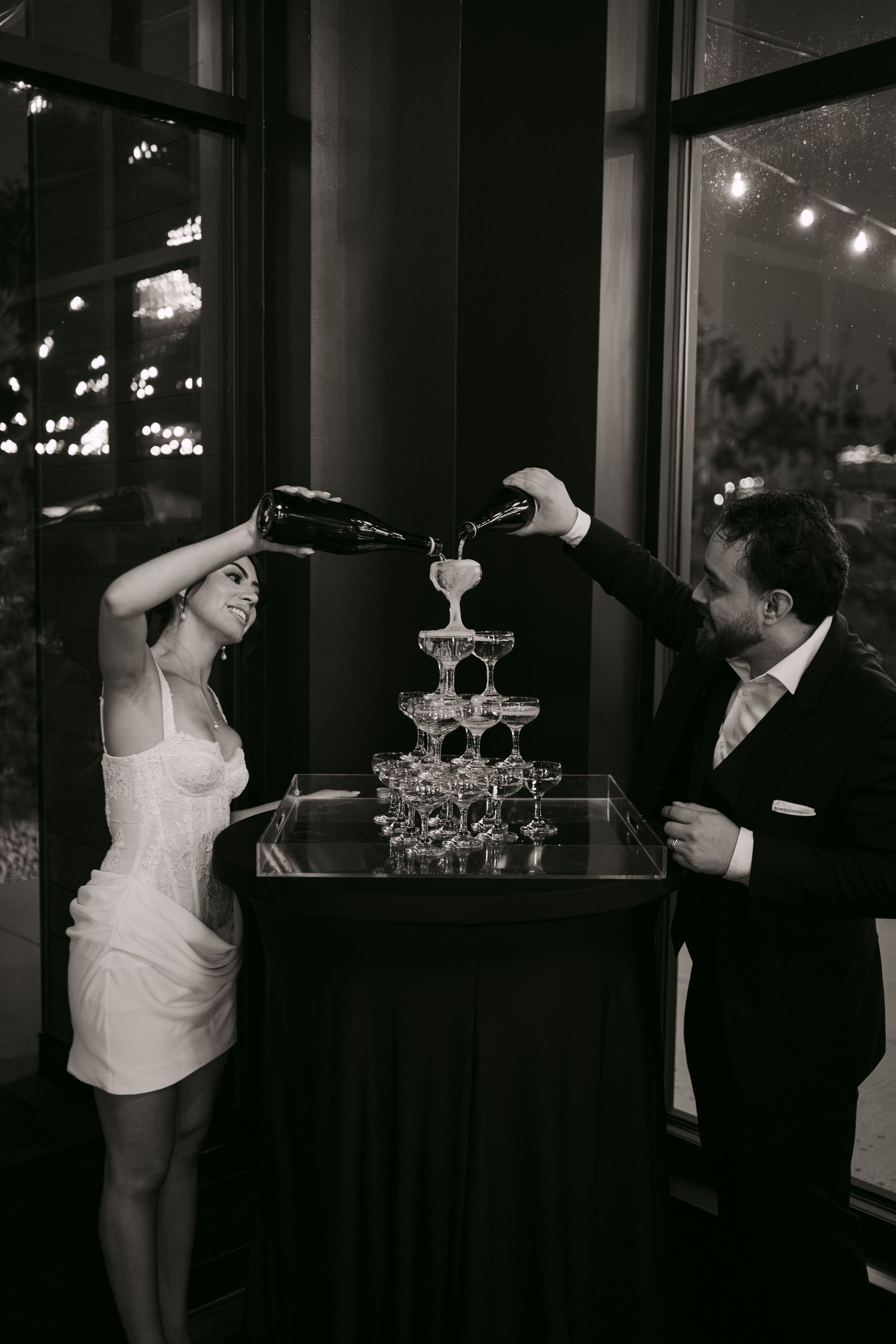 Black and white photo of bride and groom pouring champagne tower at elegant wedding reception.