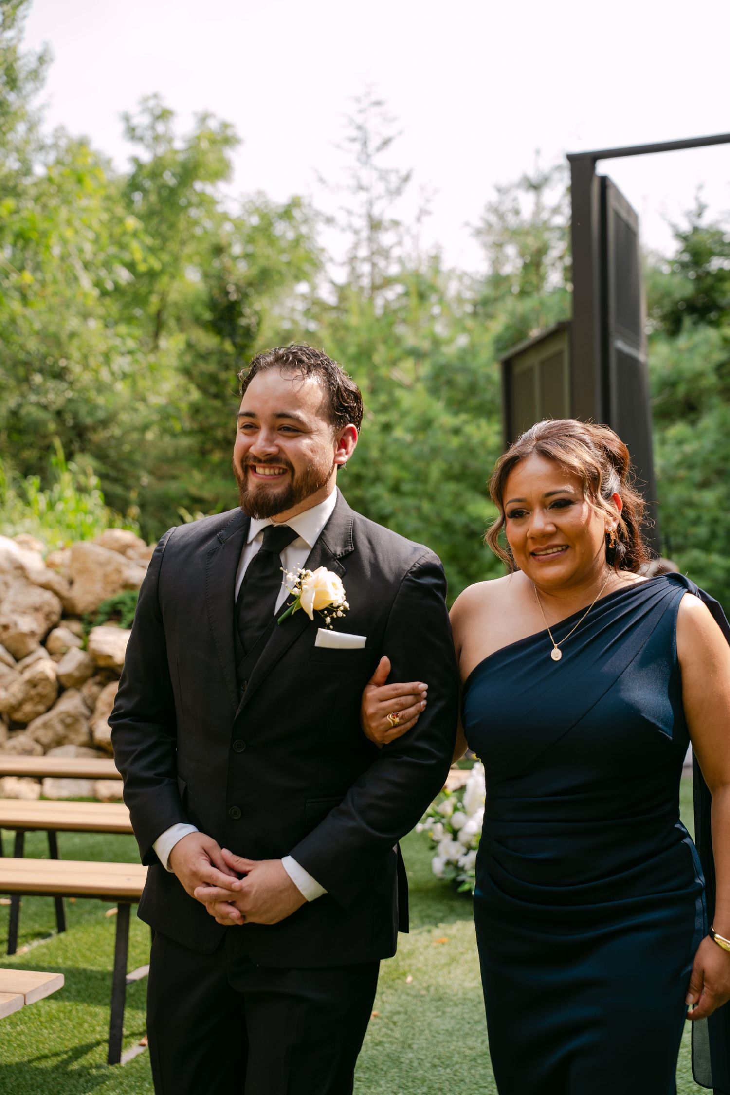 Smiling groom in black suit escorts woman in navy one-shoulder gown down outdoor wedding aisle.