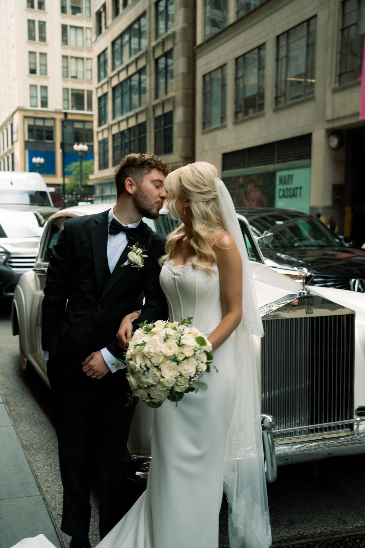 Bride and groom share a romantic moment in front of a classic white Rolls-Royce on a city street.