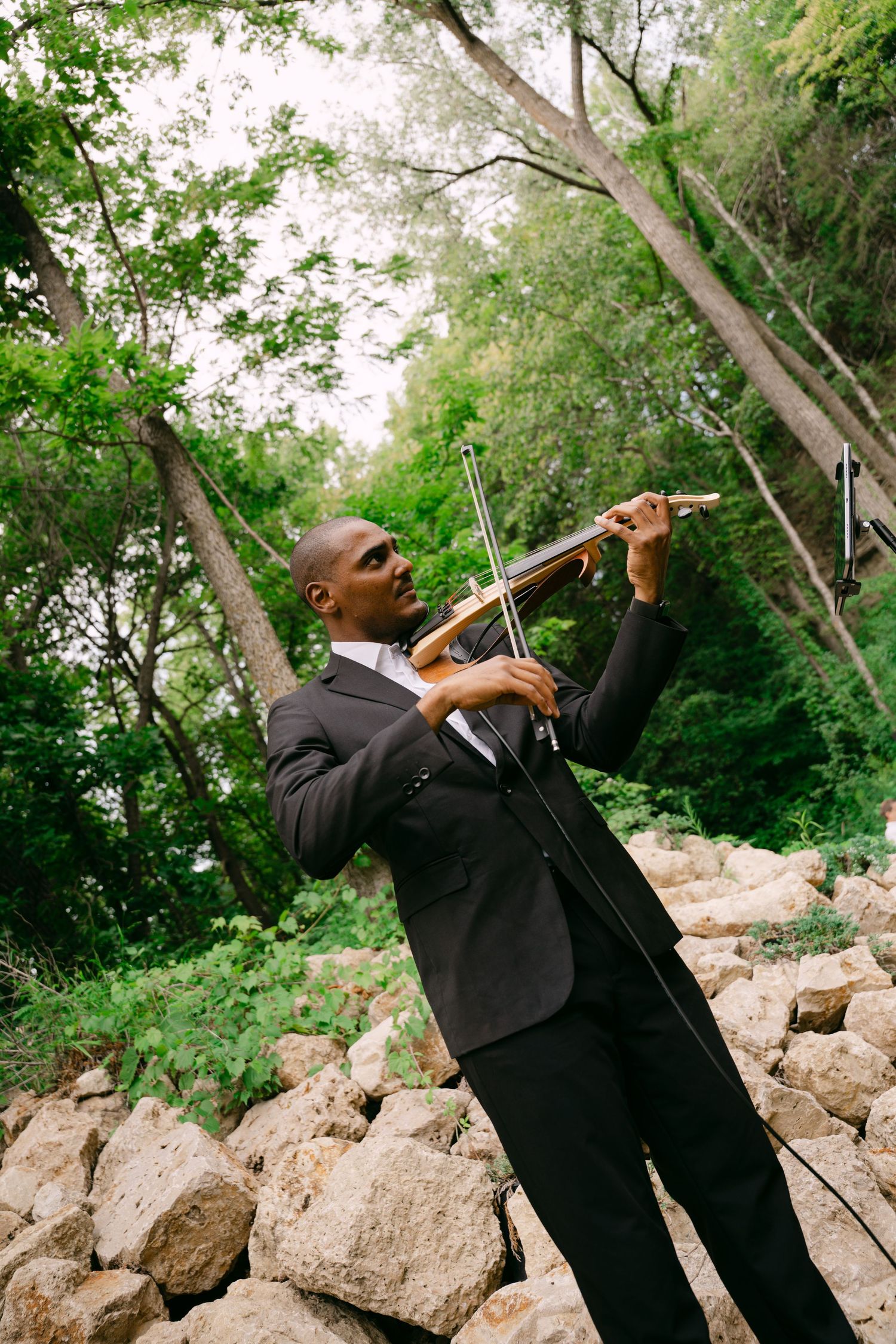 Musician in black suit playing violin outdoors among rocks and lush green trees at wedding.